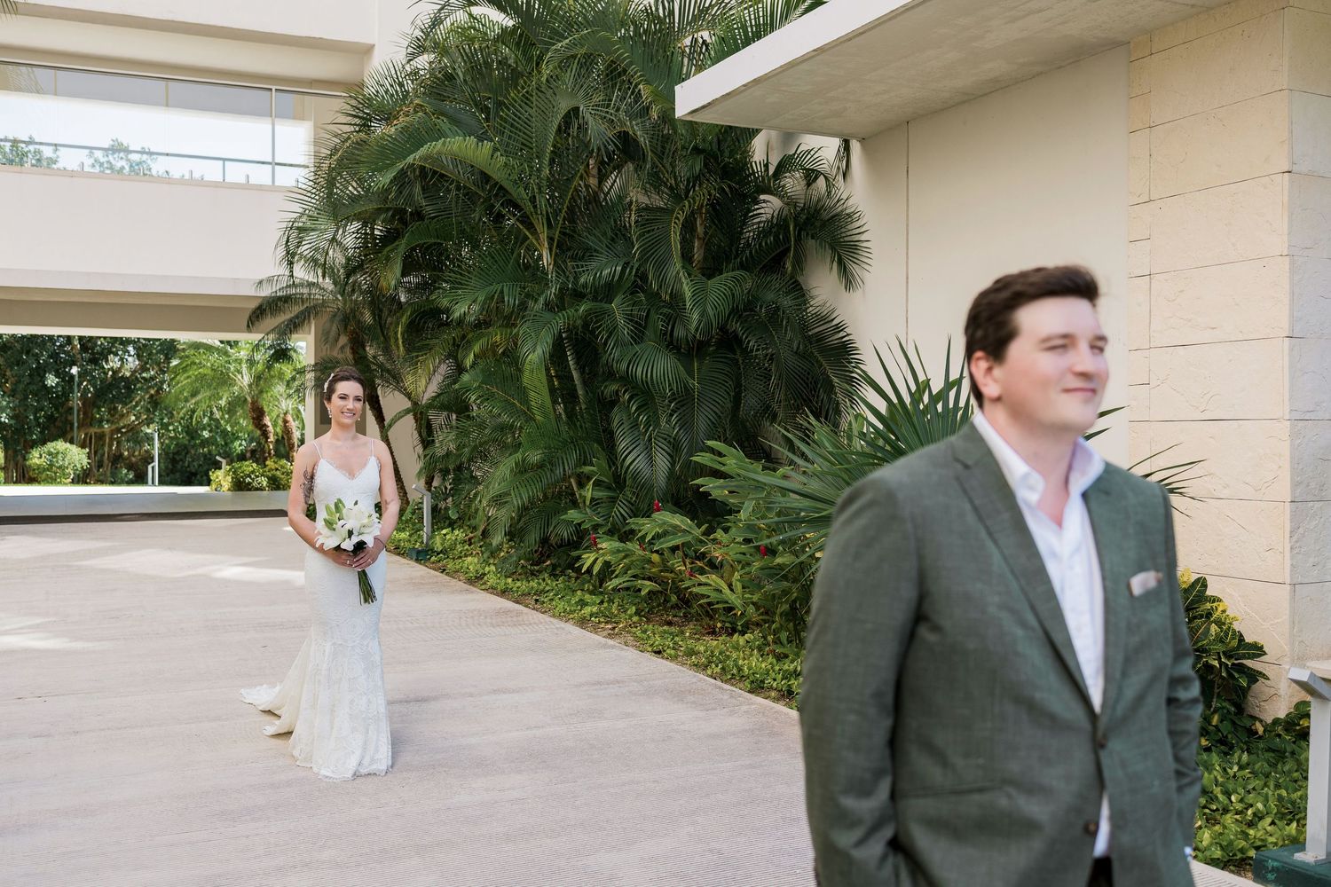 Bride in white gown approaches groom in gray suit during outdoor first look at tropical wedding venue.