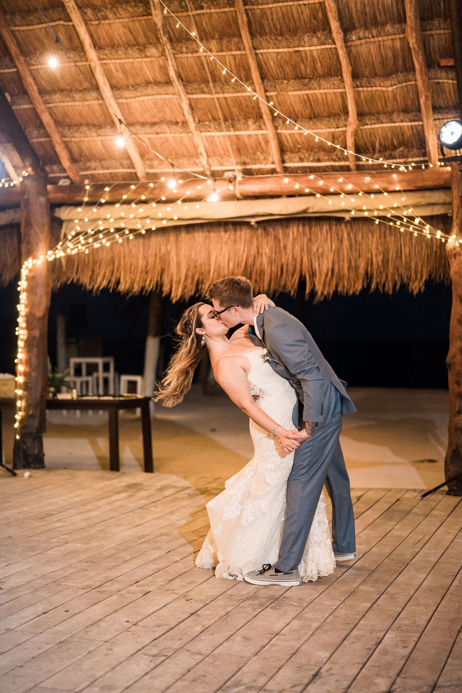 Groom dipping bride for a romantic kiss under string lights on a wooden dance floor at beach wedding.