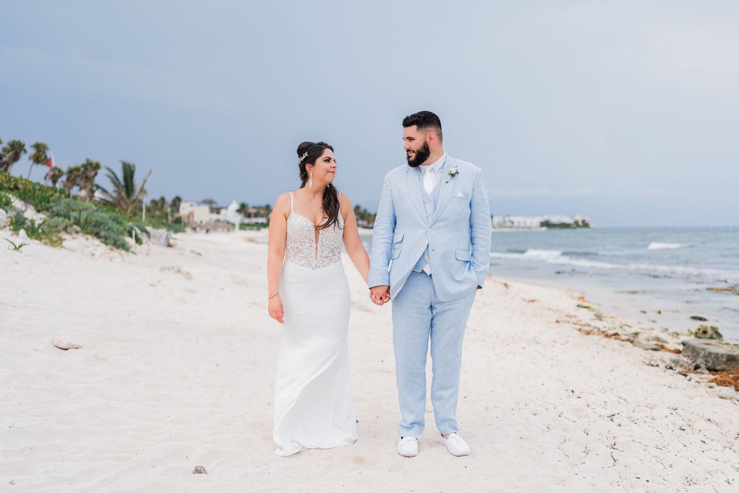Bride in white gown and bearded groom in light blue suit walk hand in hand along a sunny beach.