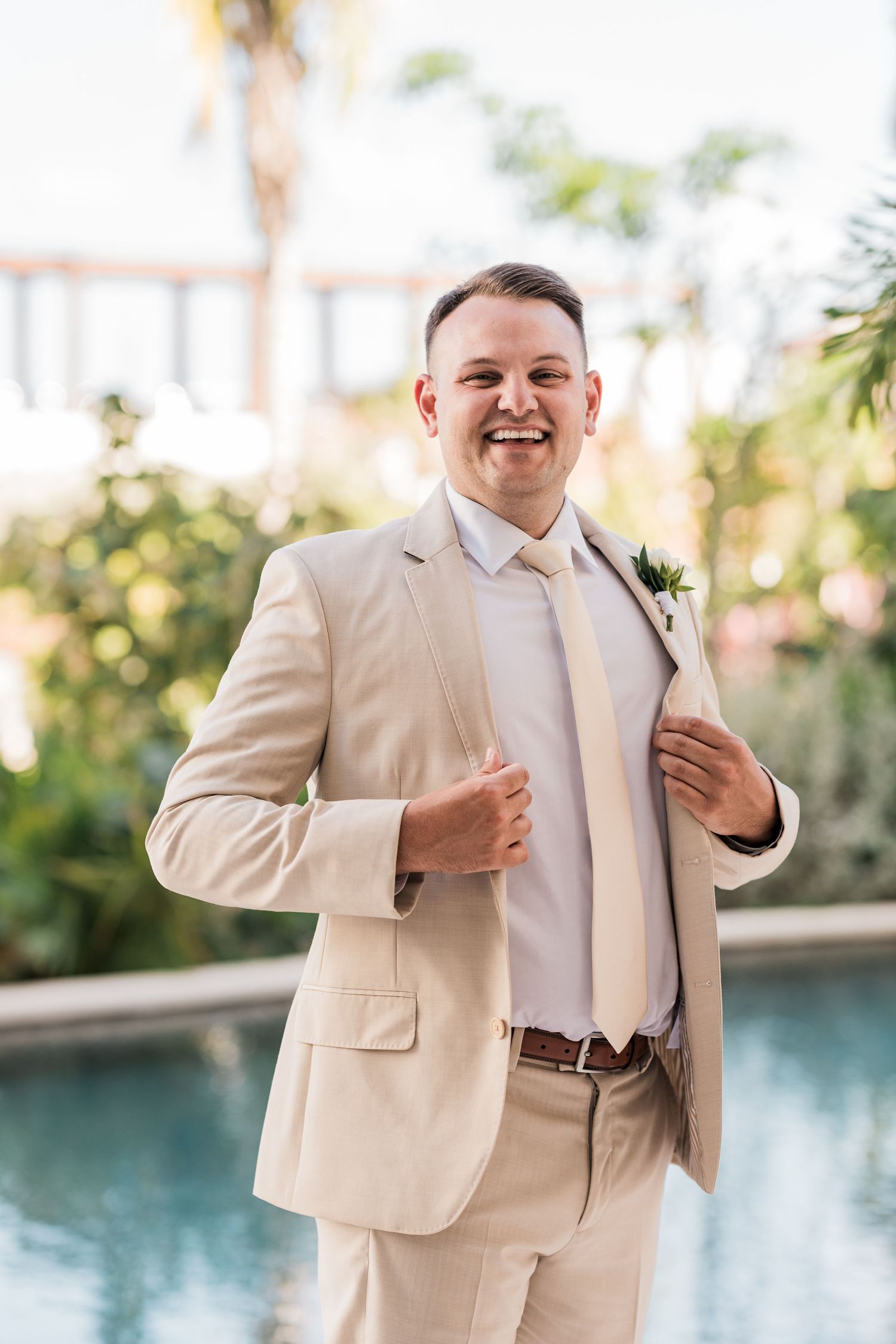 Smiling groom in tan suit with boutonniere posing confidently outdoors near a pool and palms.