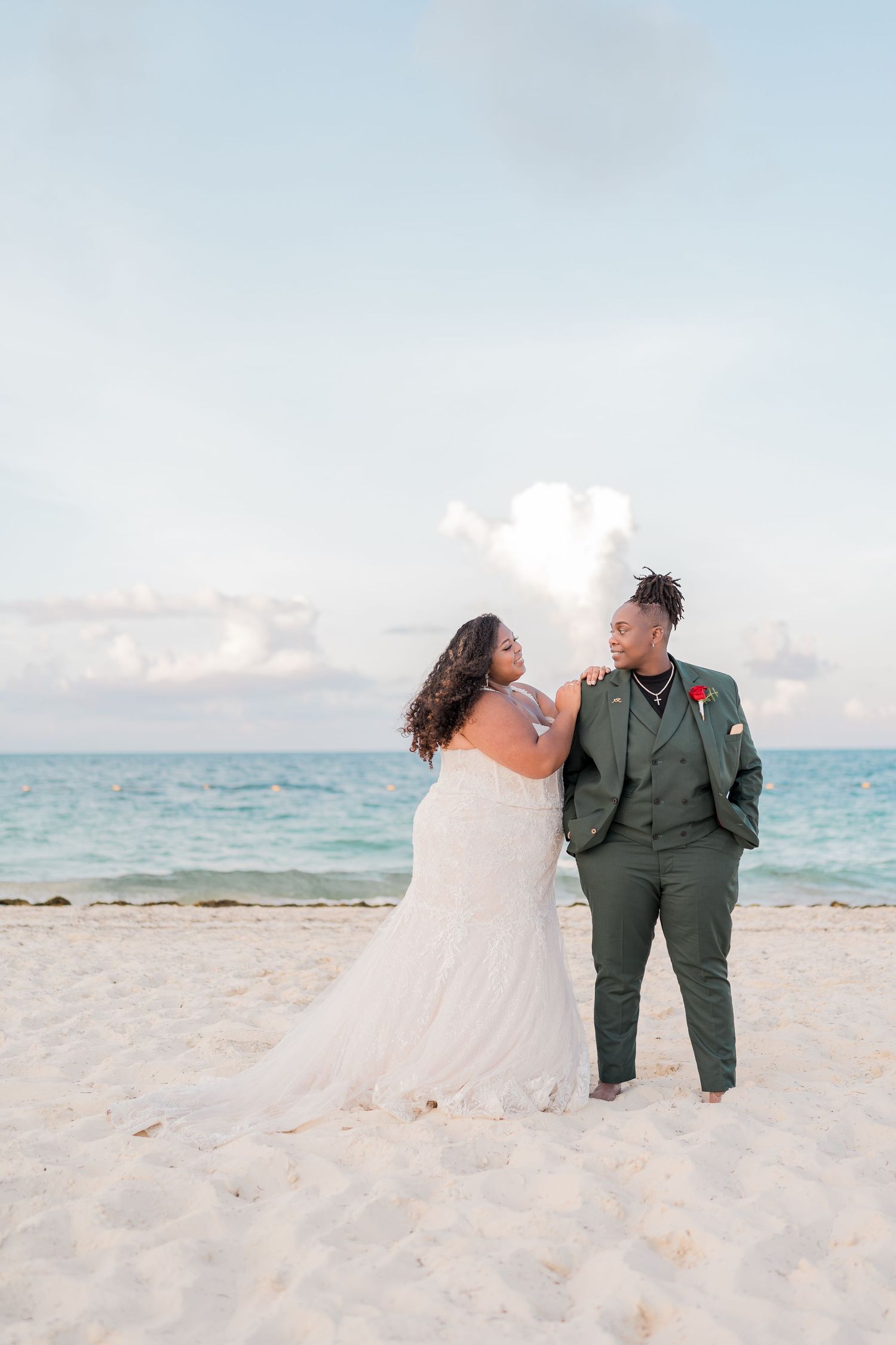 Bride in white gown and groom in green suit share a romantic moment on a white sandy Caribbean beach.