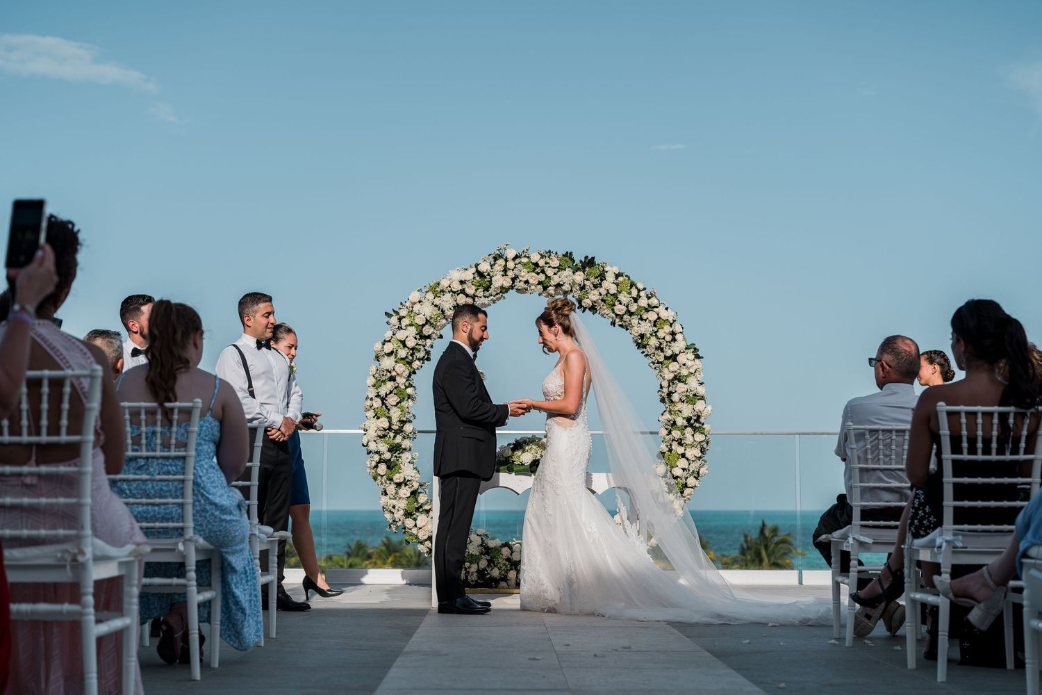 Outdoor rooftop wedding ceremony with couple at floral arch and seated guests oceanside.