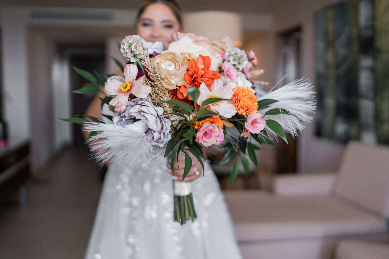Close-up of a bride holding a vibrant boho wedding bouquet with orange, pink, and white blooms and feathers.