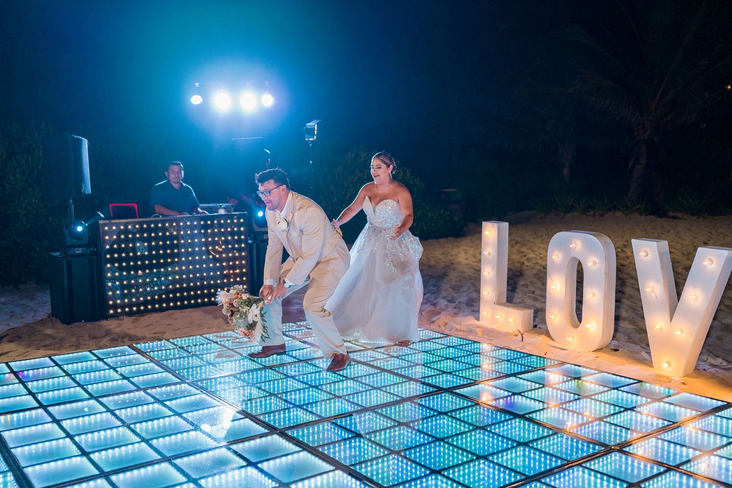 Bride and groom making grand entrance onto lit LED dance floor beside LOVE sign at beach wedding.