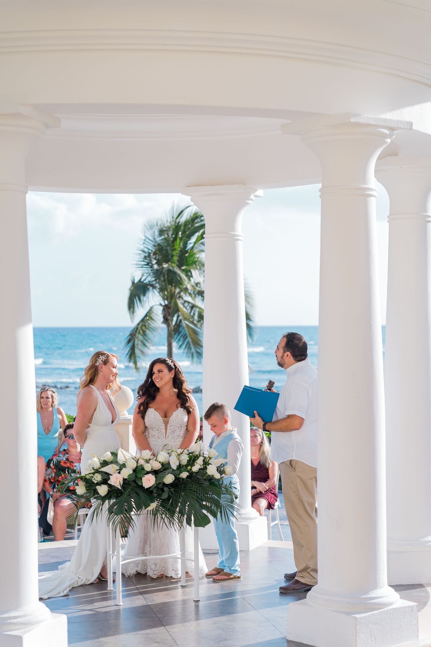 Two brides and a ring bearer at a white gazebo wedding ceremony overlooking the ocean.