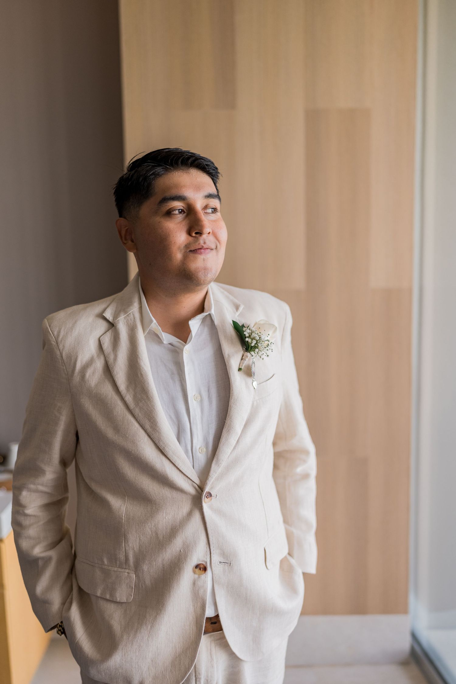 Groom in beige linen suit with floral boutonniere gazing upward before beach wedding ceremony.