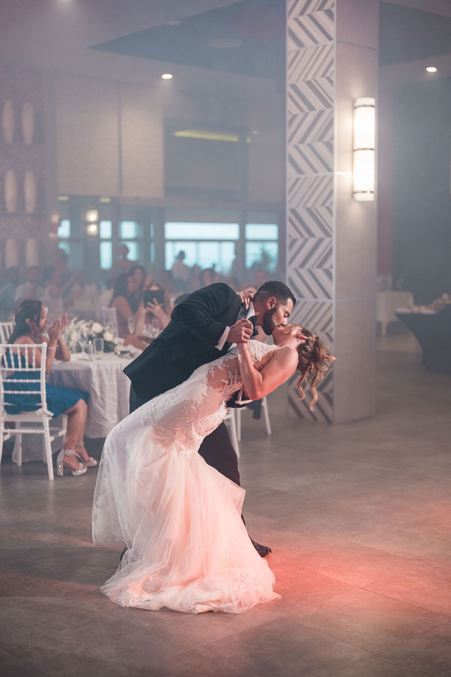 Groom dips bride dramatically during first dance in softly lit wedding reception with guests watching.
