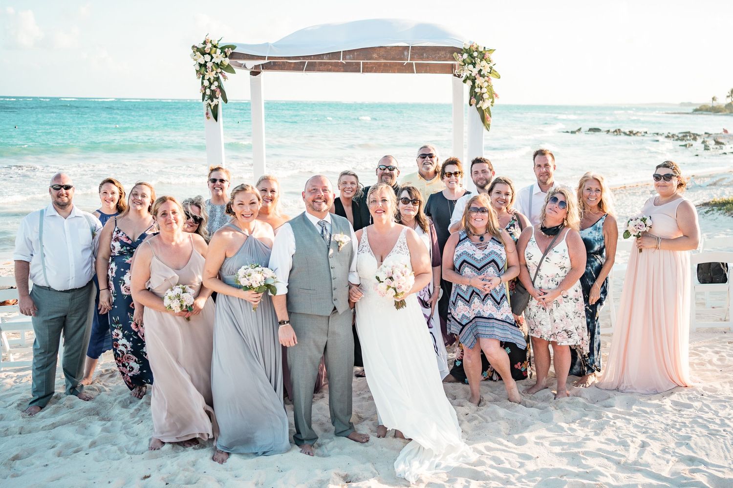 Wedding party and guests pose together under floral arch on tropical beach after ceremony.