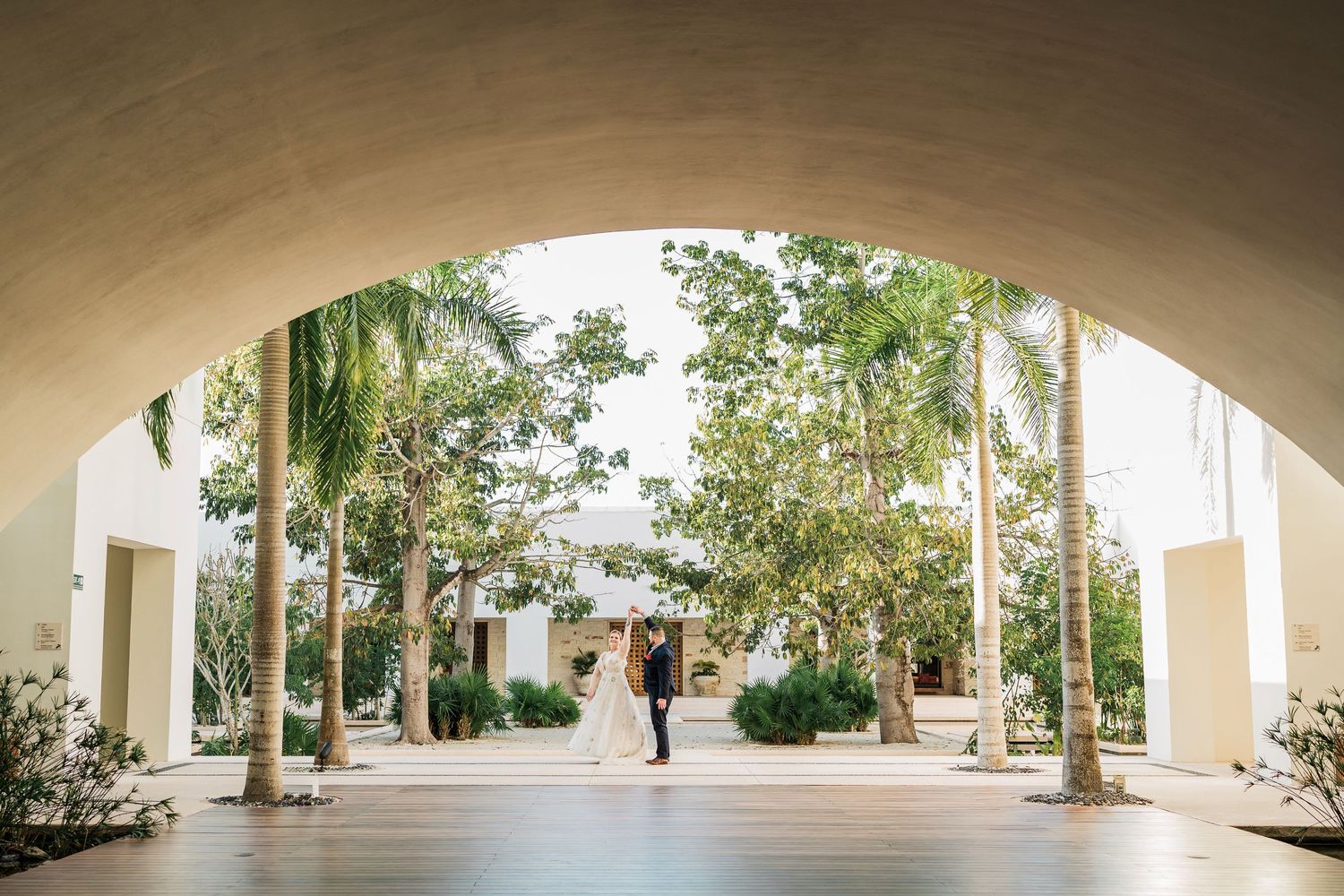 Secrets Akumal Wedding - Couple stands under elegant arched corridor surrounded by palm trees at a sunlit outdoor wedding venue.