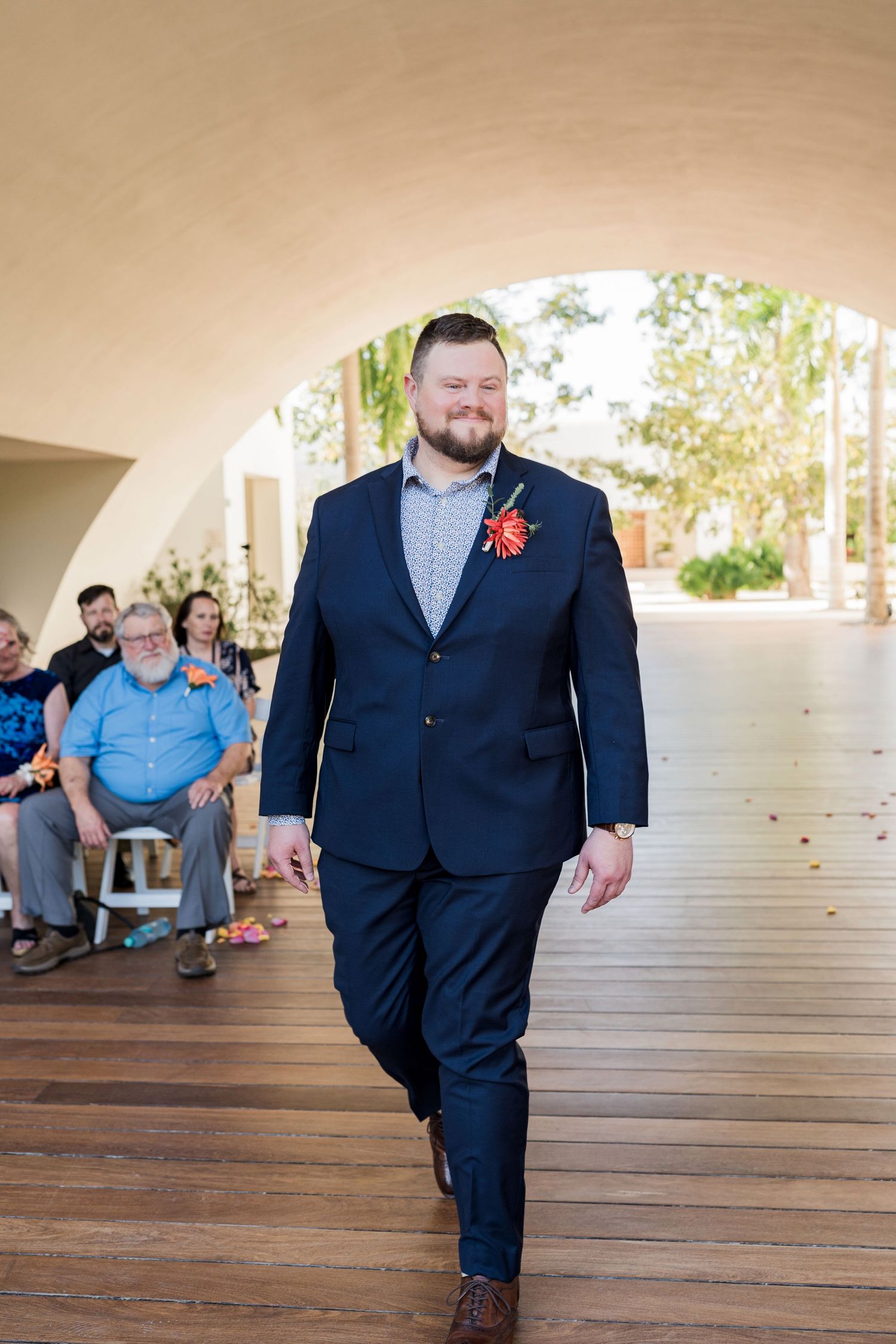 Secrets Akumal Wedding - Groom in navy blue suit with red boutonniere walks down aisle at outdoor wedding ceremony.