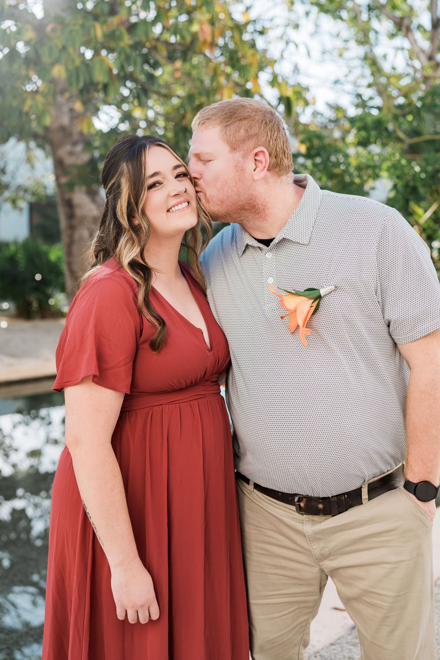 Secrets Akumal Wedding -A couple poses outdoors, man kissing woman's cheek, she wears red dress, he wears gray polo with orange boutonniere.