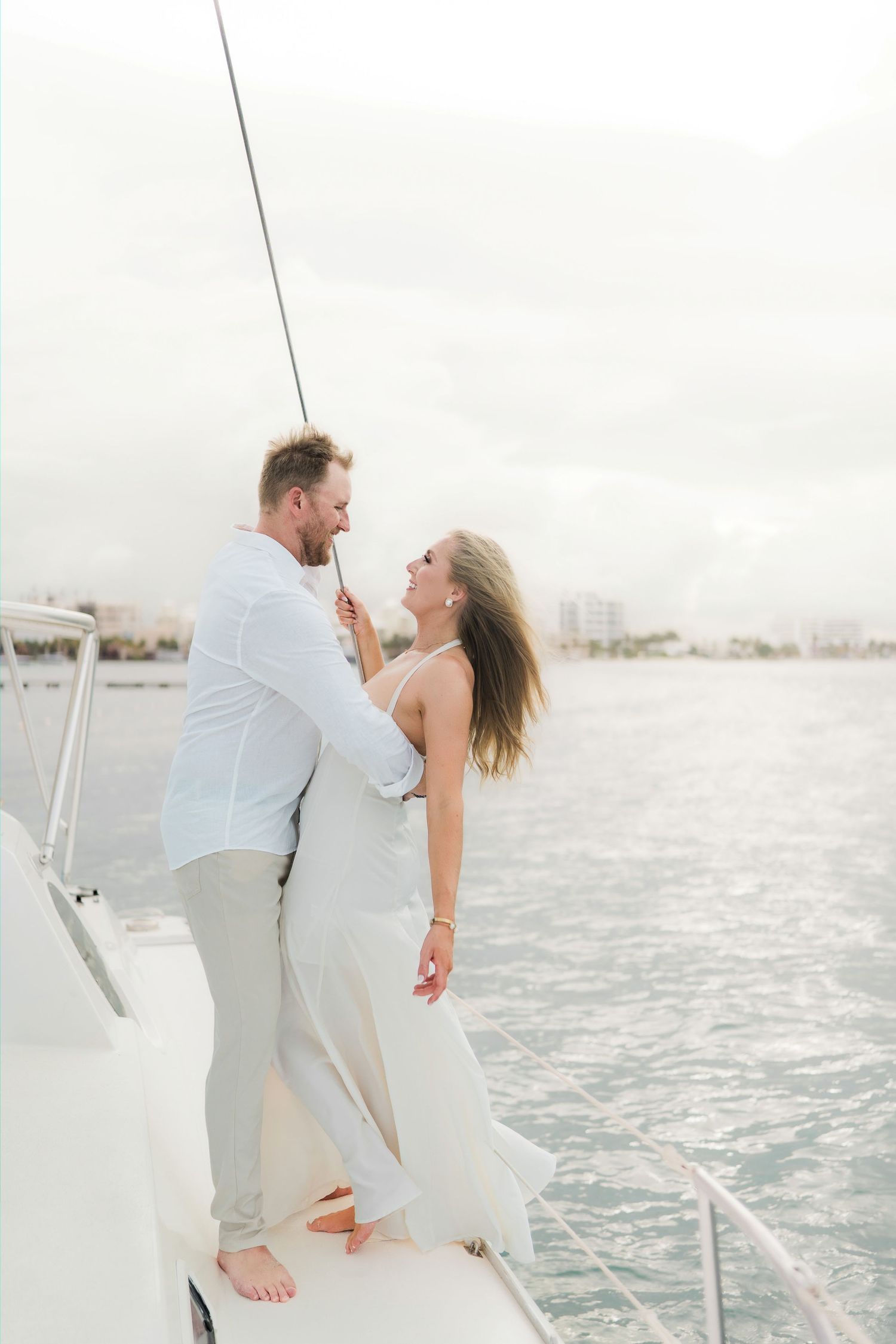 Cancun Engagement Photoshoot - Couple in white outfits embracing on a yacht deck with ocean and city skyline in the background.