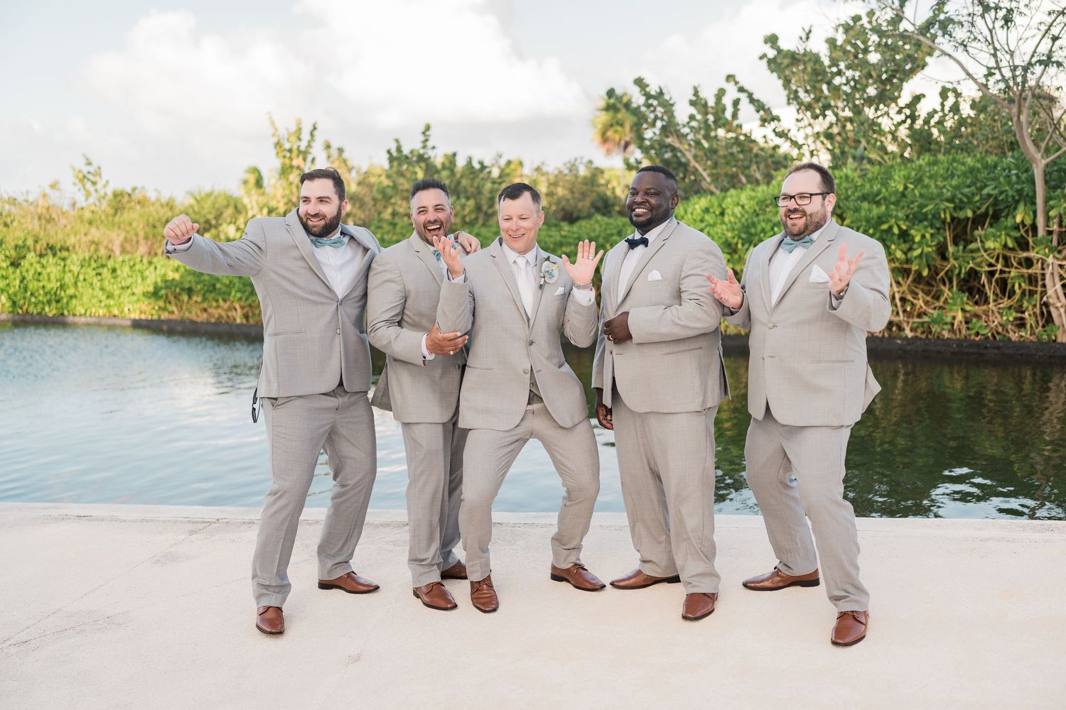 Five groomsmen in light gray suits pose playfully near a waterway at grand palladium costa mujeres wedding