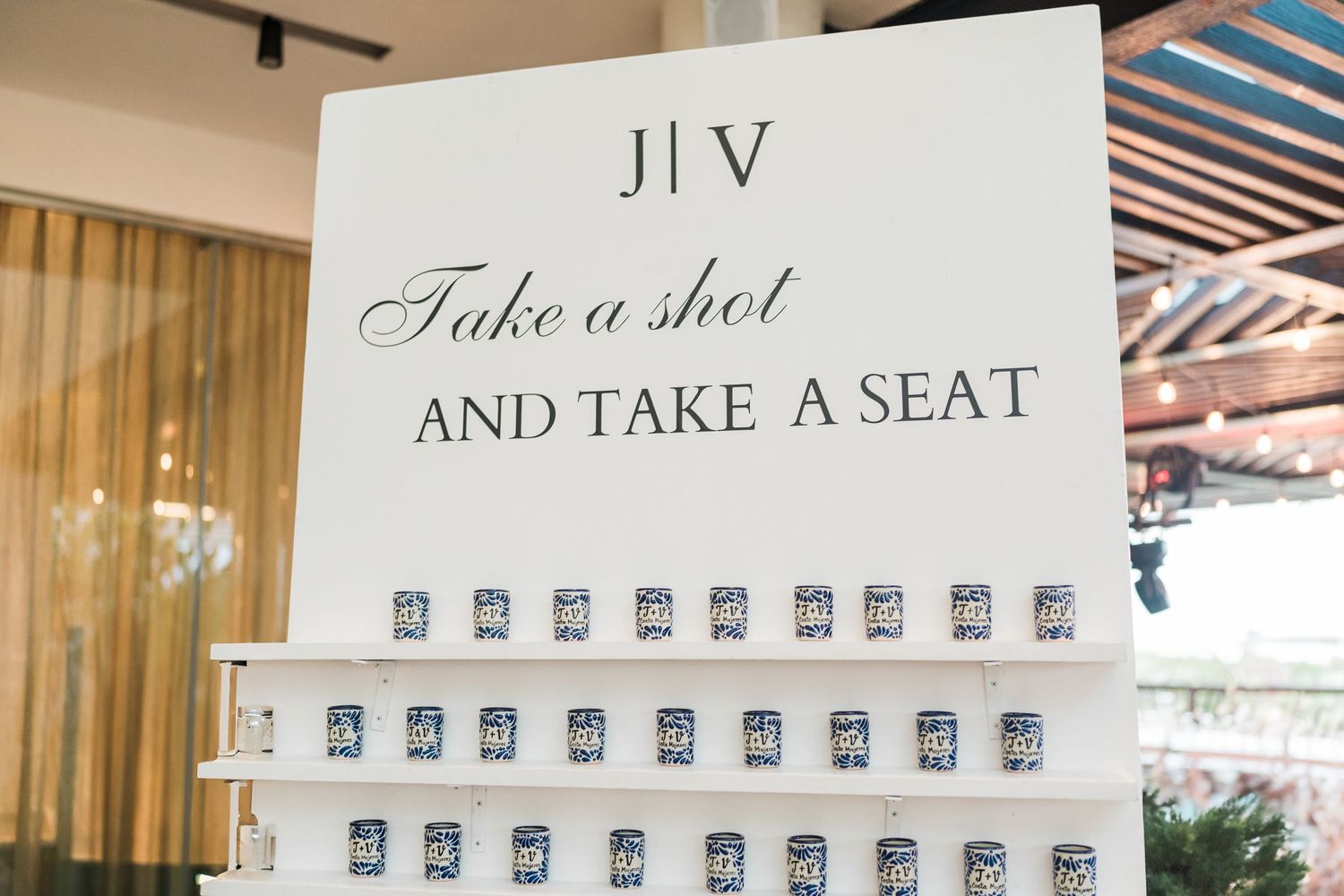Wedding seating chart sign reading Take a Shot and Take a Seat with mini shot glasses displayed on white shelves.