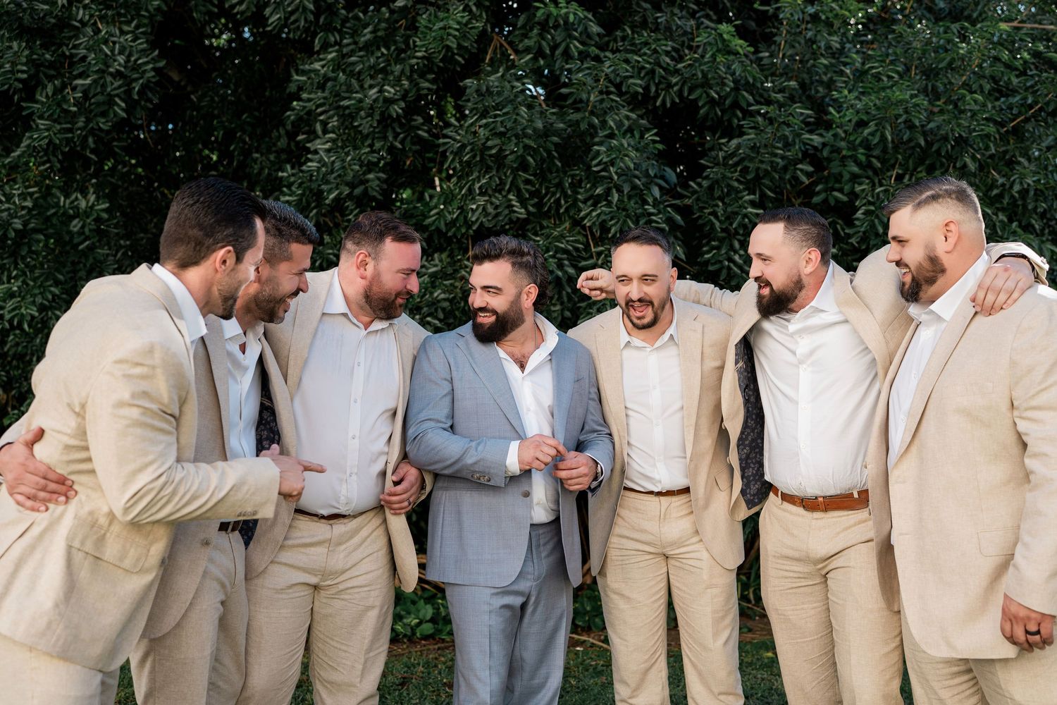 Groom in gray suit laughing with groomsmen in tan suits outdoors near lush green trees at a wedding in Hard Rock Riviera Maya