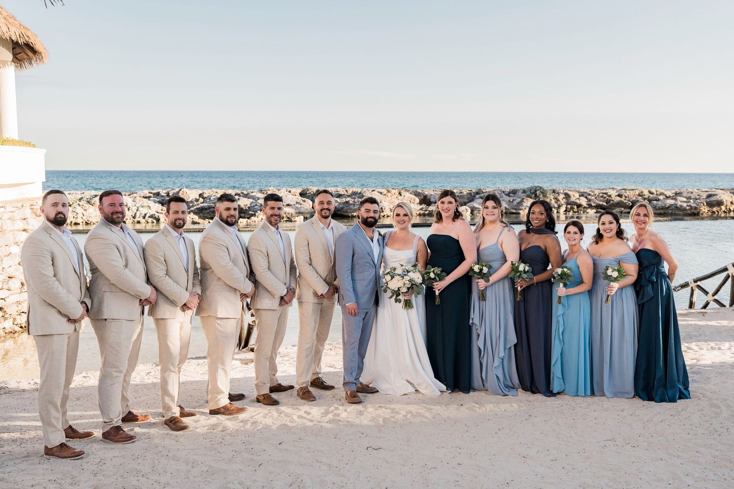 Wedding party poses on beach, groomsmen in tan suits and bridesmaids in blue gowns with bride and groom centered.