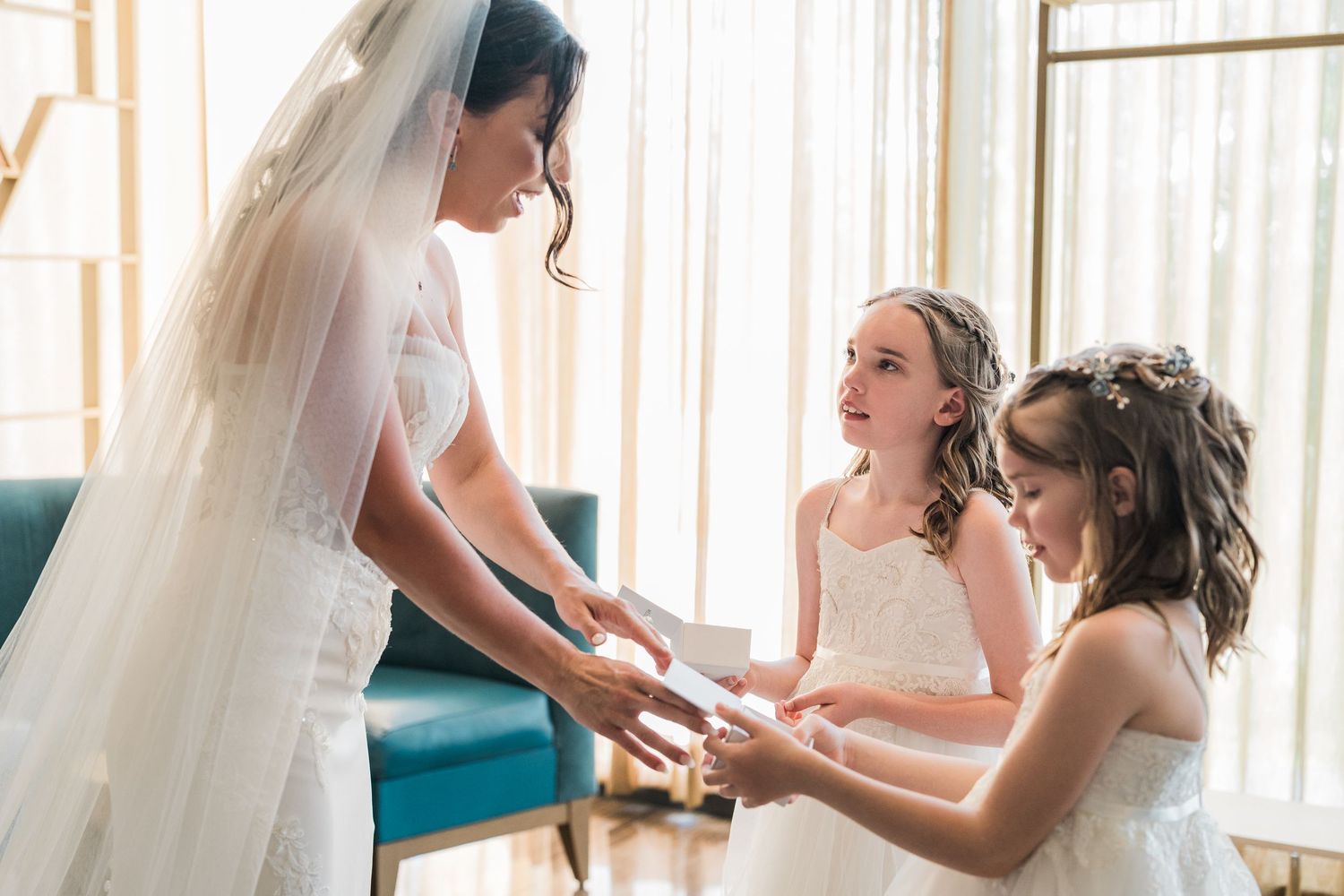 Bride in white wedding gown and veil holding hands with two young flower girls in white dresses near bright windows.