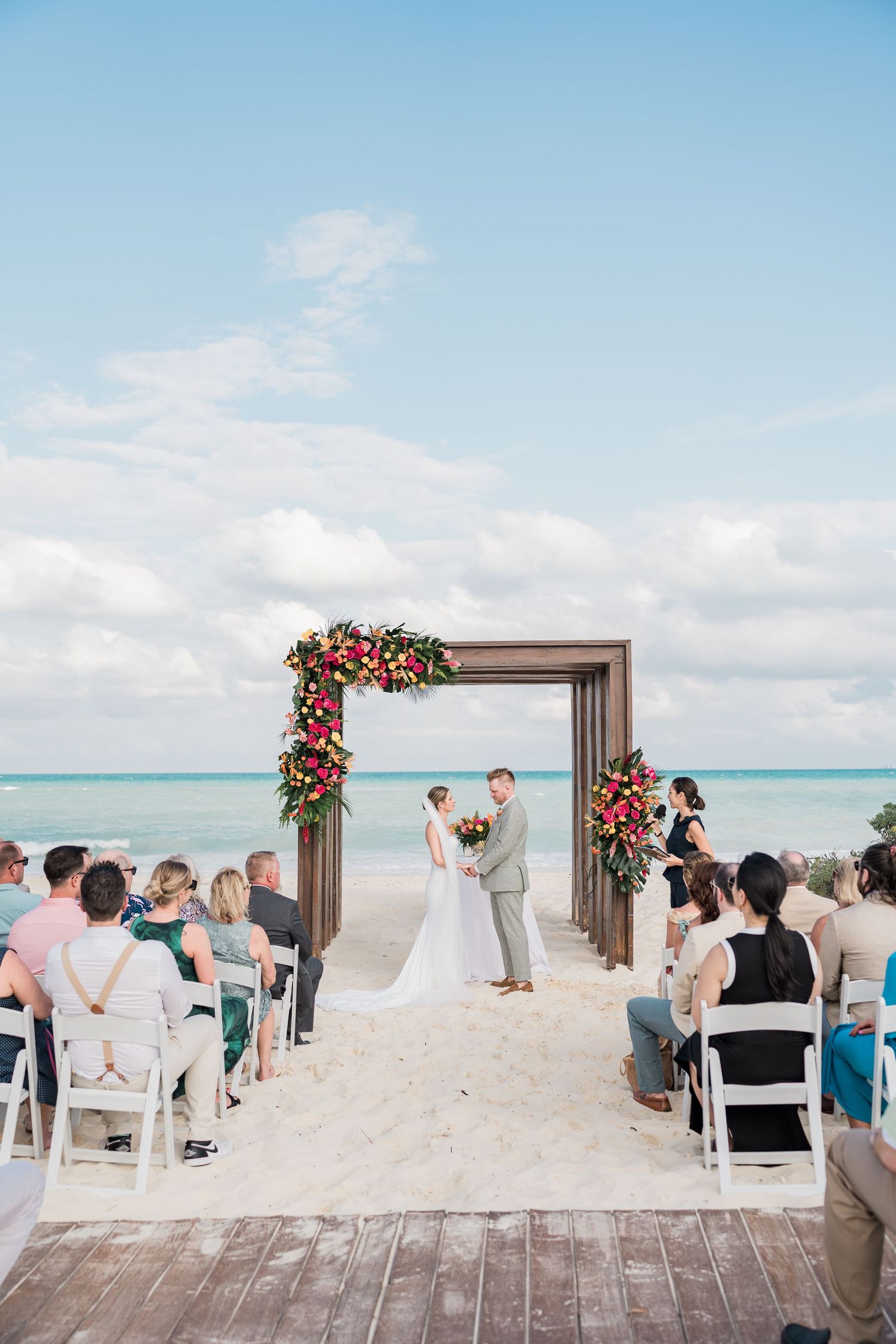 Romantic beach wedding ceremony with floral arch, ocean backdrop, and guests seated on white chairs.