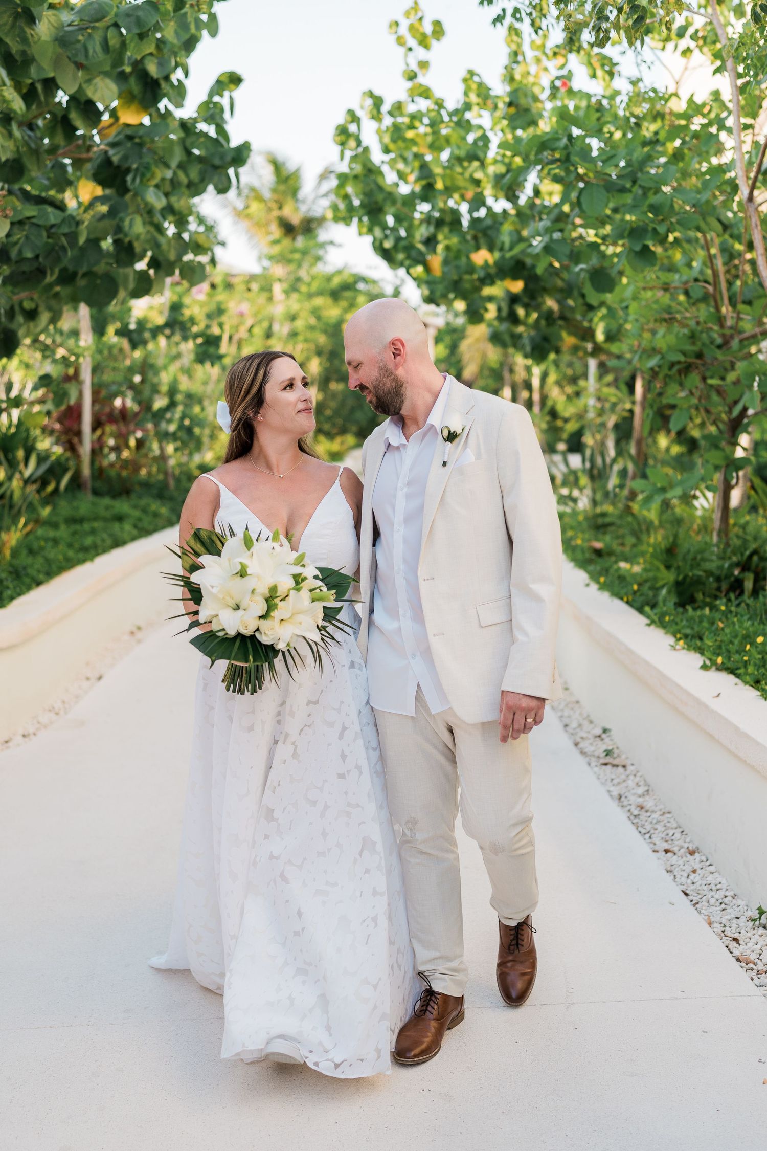 Bride and groom walk hand in hand on a tropical path, she holds white florals, he wears a tan suit.