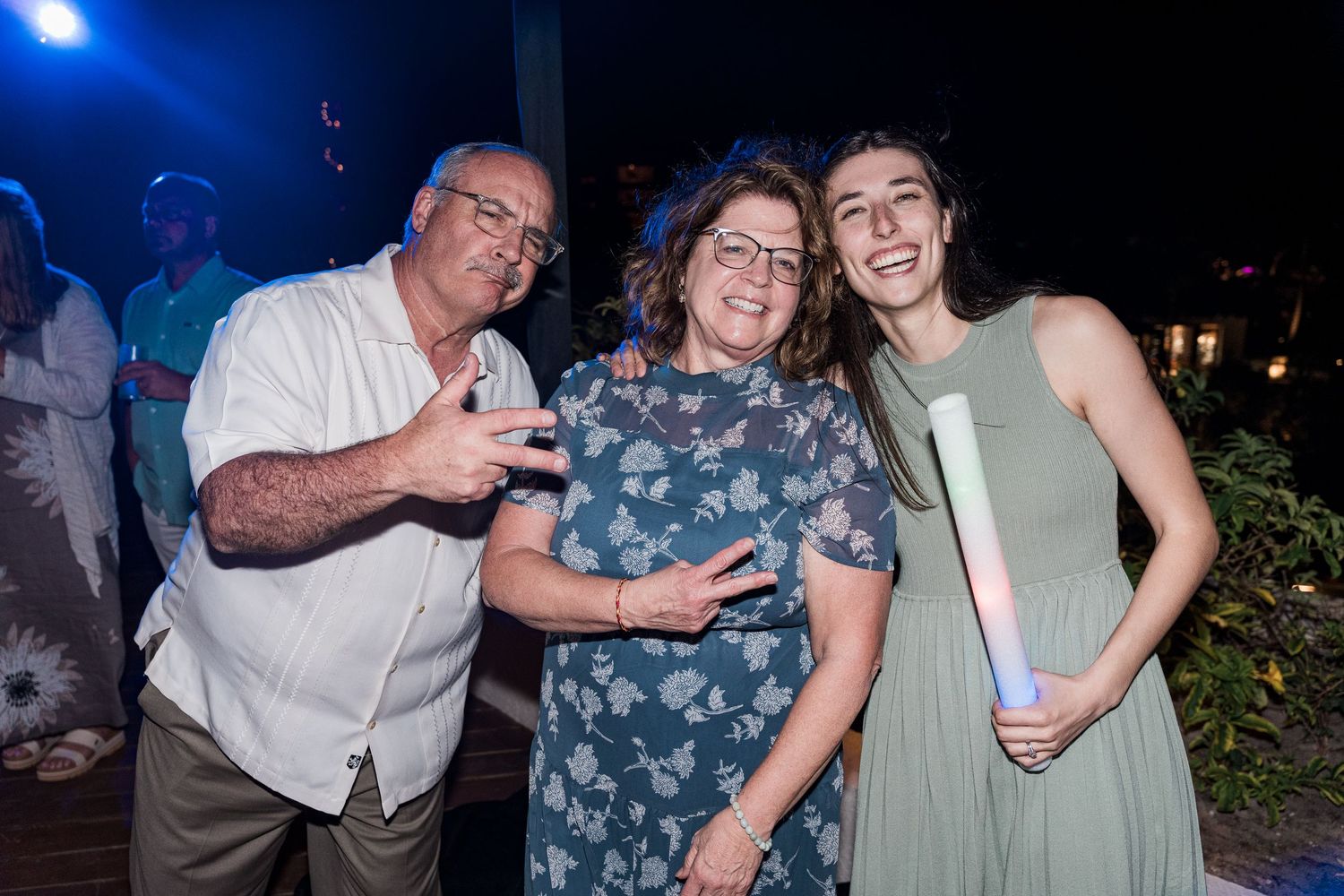 Three people smiling and posing together at an outdoor evening party, with festive lighting in the background.