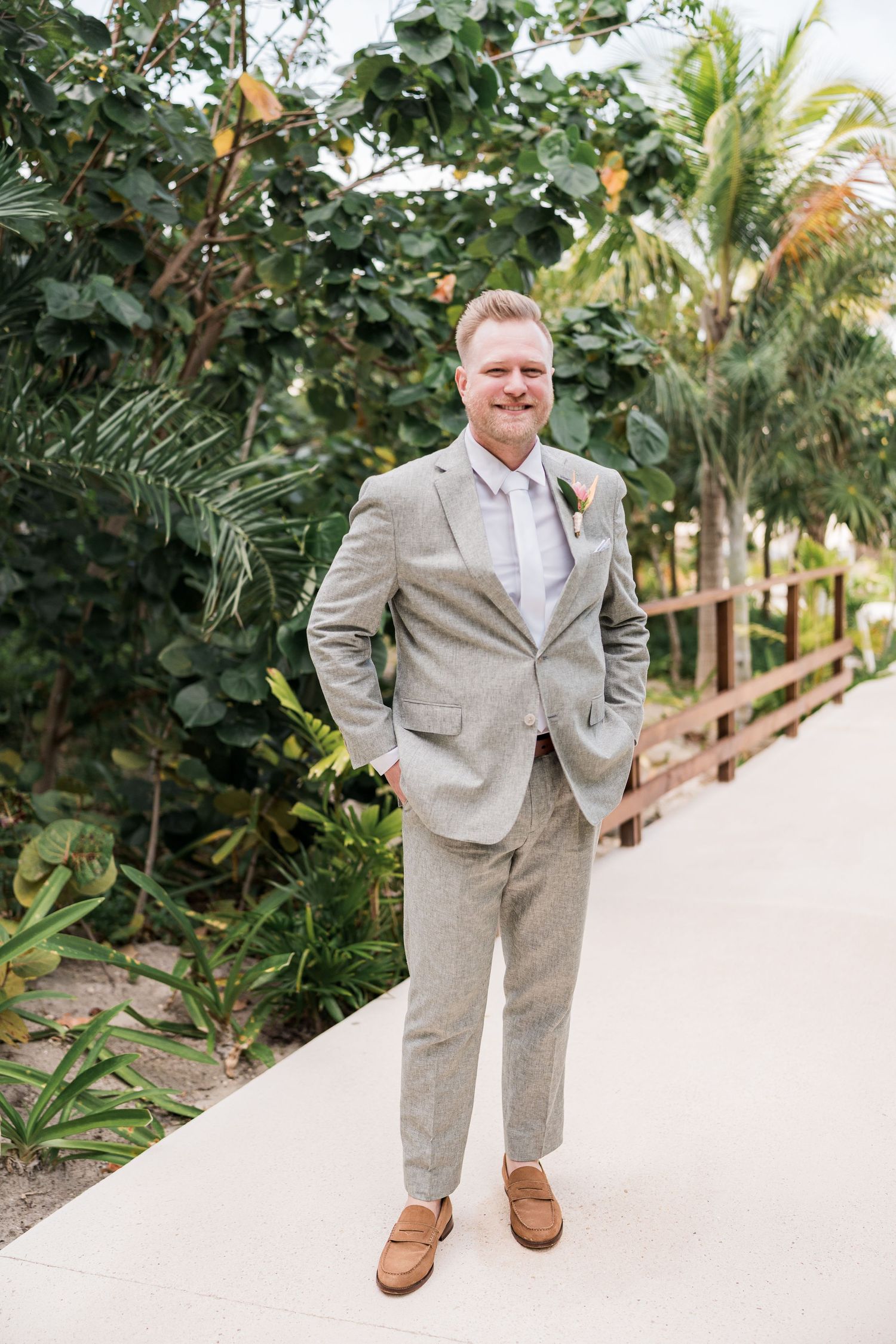 Groom in light gray linen suit and brown loafers smiling outdoors on a tropical wedding day.