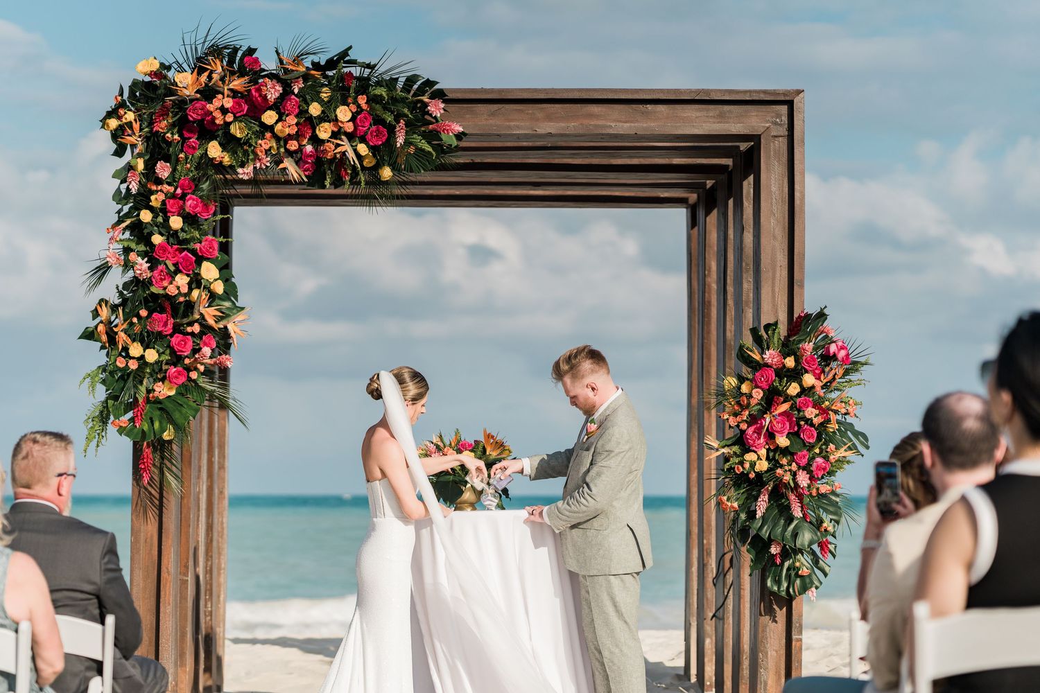 Couple exchanging vows at a beach wedding ceremony under a wooden arch adorned with vibrant tropical flowers.