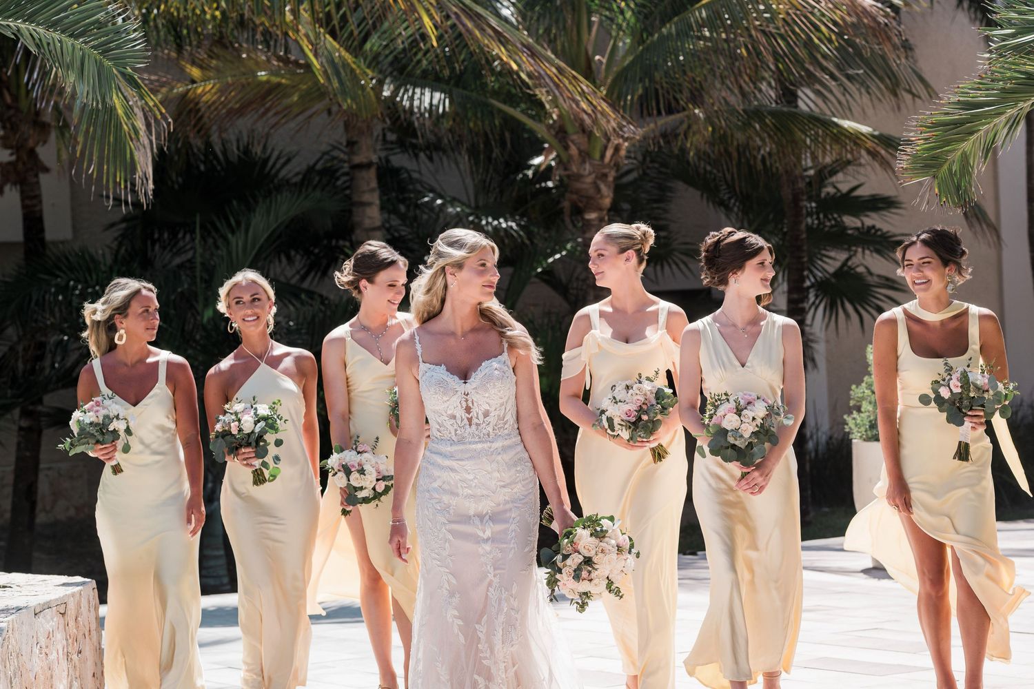 Bride in lace gown walks with six bridesmaids in yellow dresses holding bouquets near tropical palm trees.