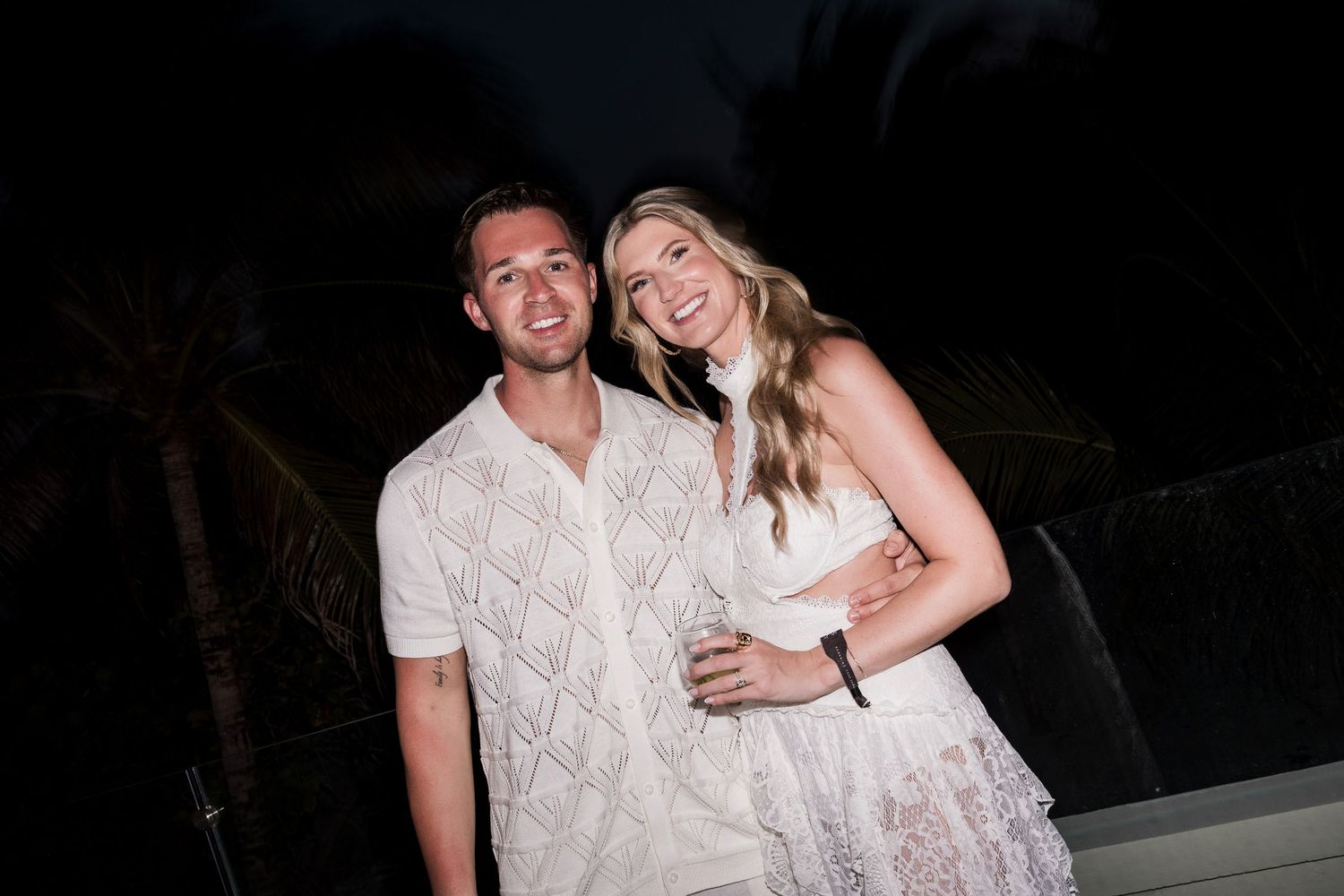 A smiling couple dressed in white poses together outdoors at night with tropical palm trees in the background.