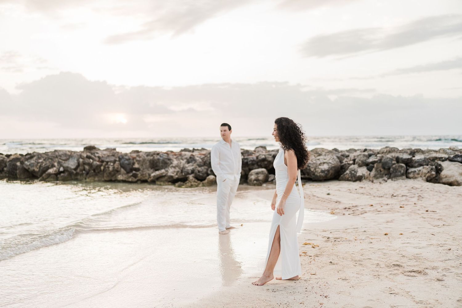 Couple in white outfits standing on a sandy beach at sunset with rocky shoreline in the background.