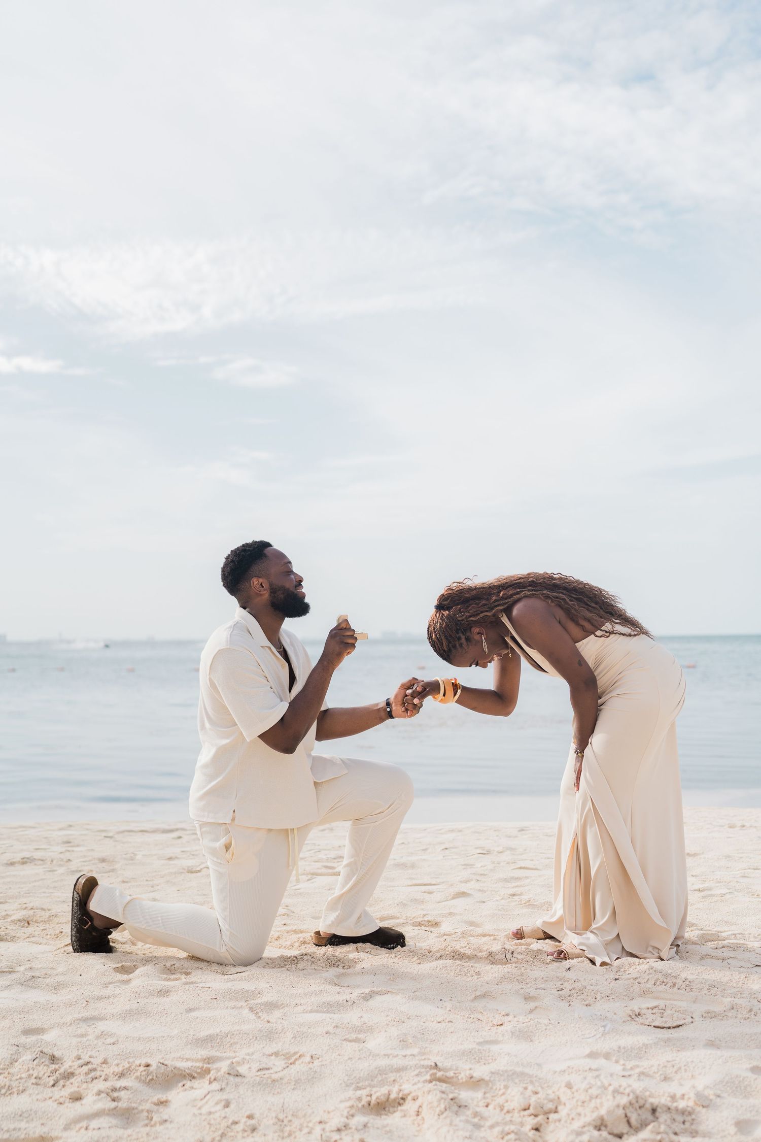 A man proposes on one knee to a woman on a sandy beach, both dressed in elegant cream outfits with the ocean behind them.