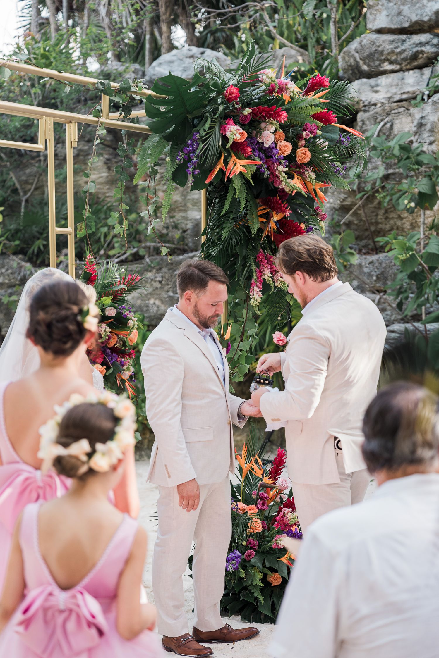 Two grooms exchange vows at tropical outdoor wedding ceremony with vibrant floral arch of birds of paradise and roses.