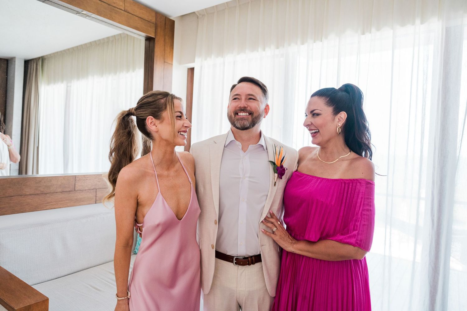 A smiling groom in beige suit stands between two women in pink dresses at a bright, elegant wedding venue.