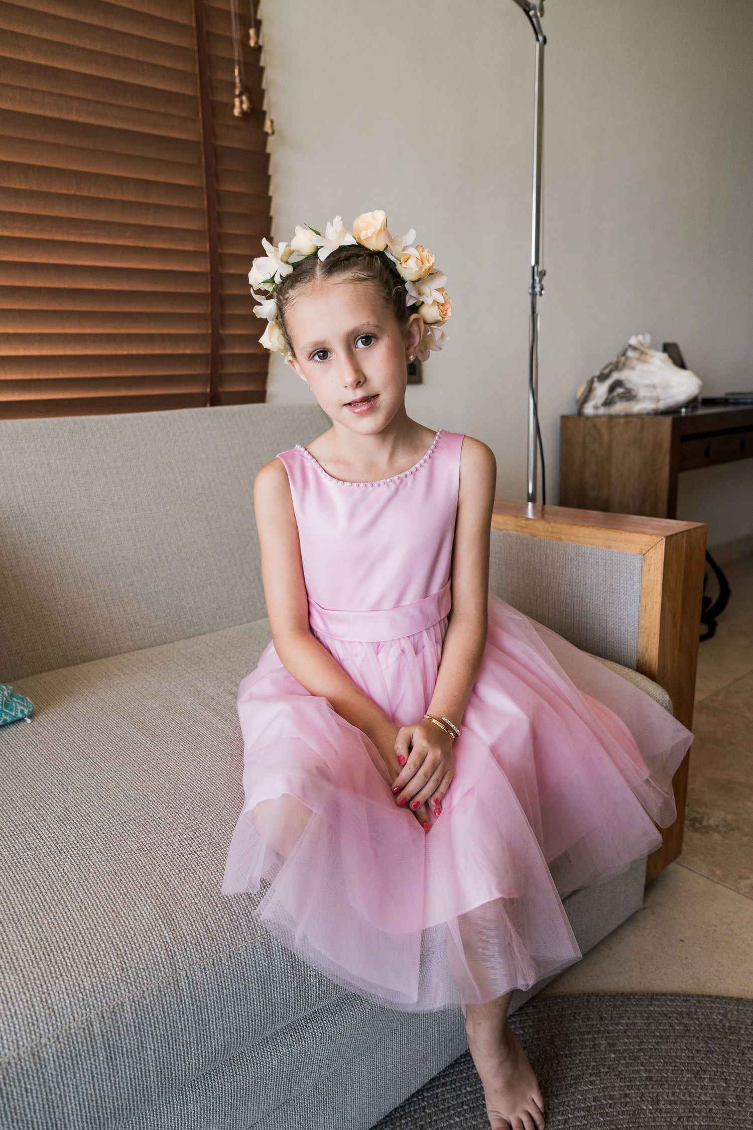 Young girl in pink tulle dress and floral crown sits gracefully on a sofa, perfect for flower girl fashion inspiration.
