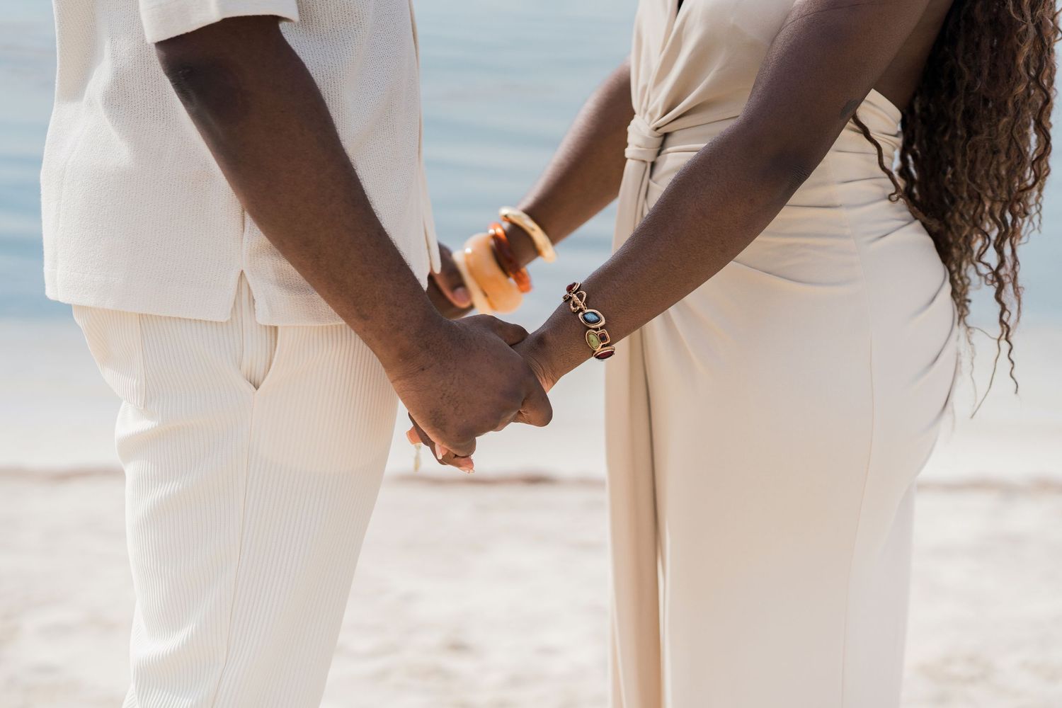 Close-up of a couple holding hands on the beach, dressed in matching white outfits with gold jewelry accents.
