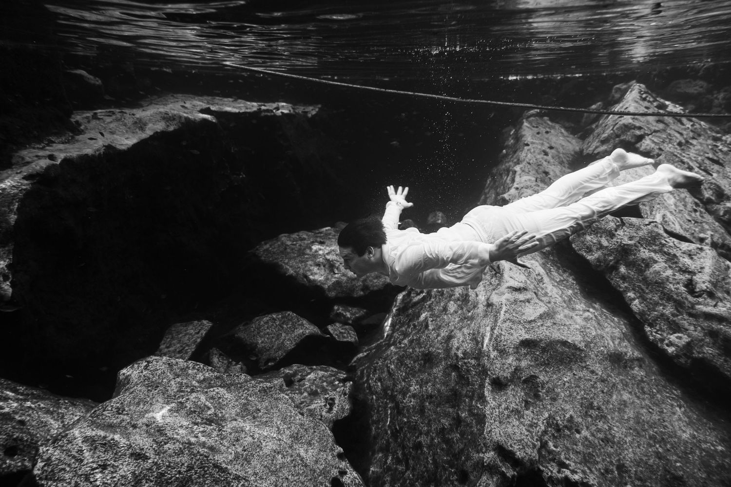 Black and white underwater photo of a person in white dress reaching upward among dark rocks.