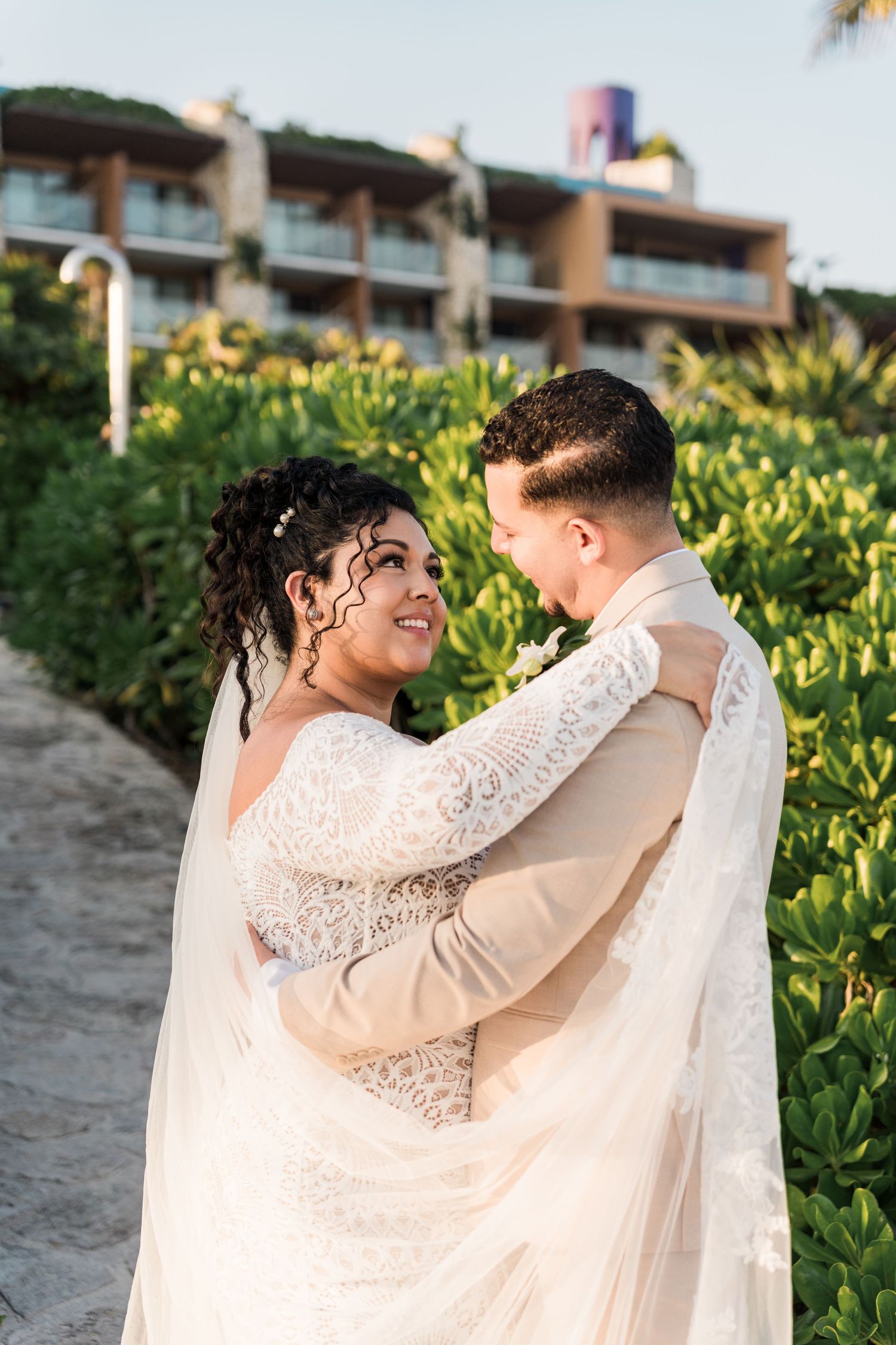 Xcaret Arte Wedding - Bride and groom embrace outdoors near lush greenery, smiling at each other in wedding attire