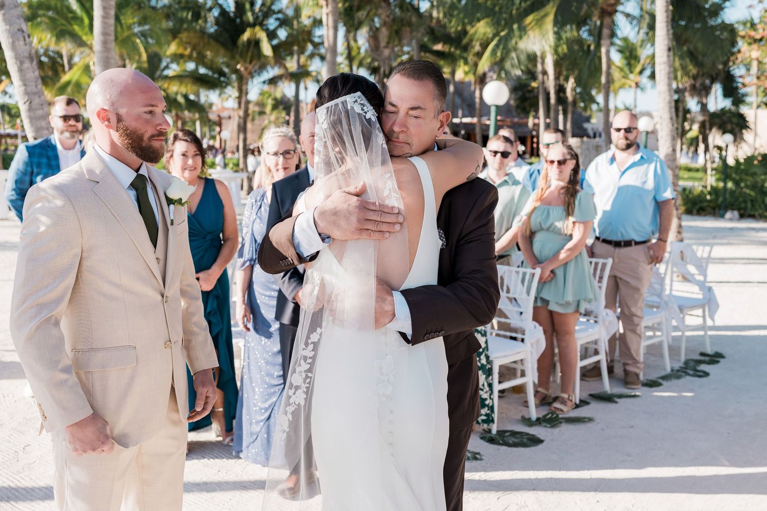 Bride and groom share an emotional hug during outdoor beach wedding ceremony with guests seated in white chairs.