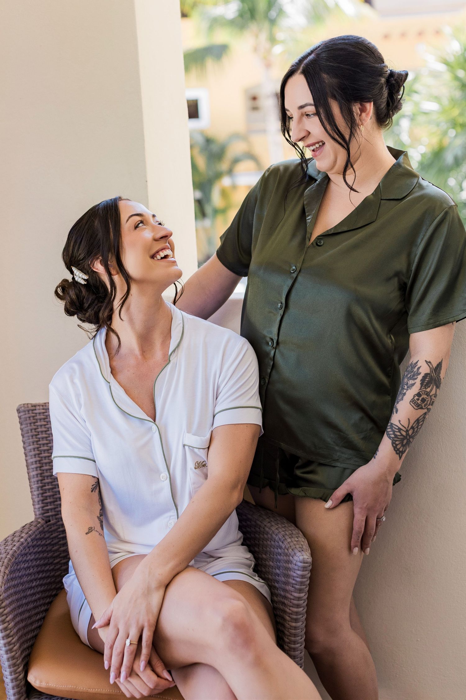 Two smiling women in satin pajamas share a joyful moment on a sunny balcony, one seated and one standing with tattoos.