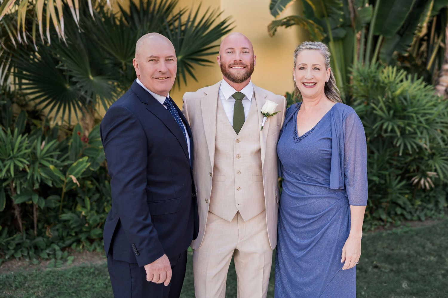 Groom in beige suit with two guests in navy attire smiling outdoors at a tropical wedding venue.