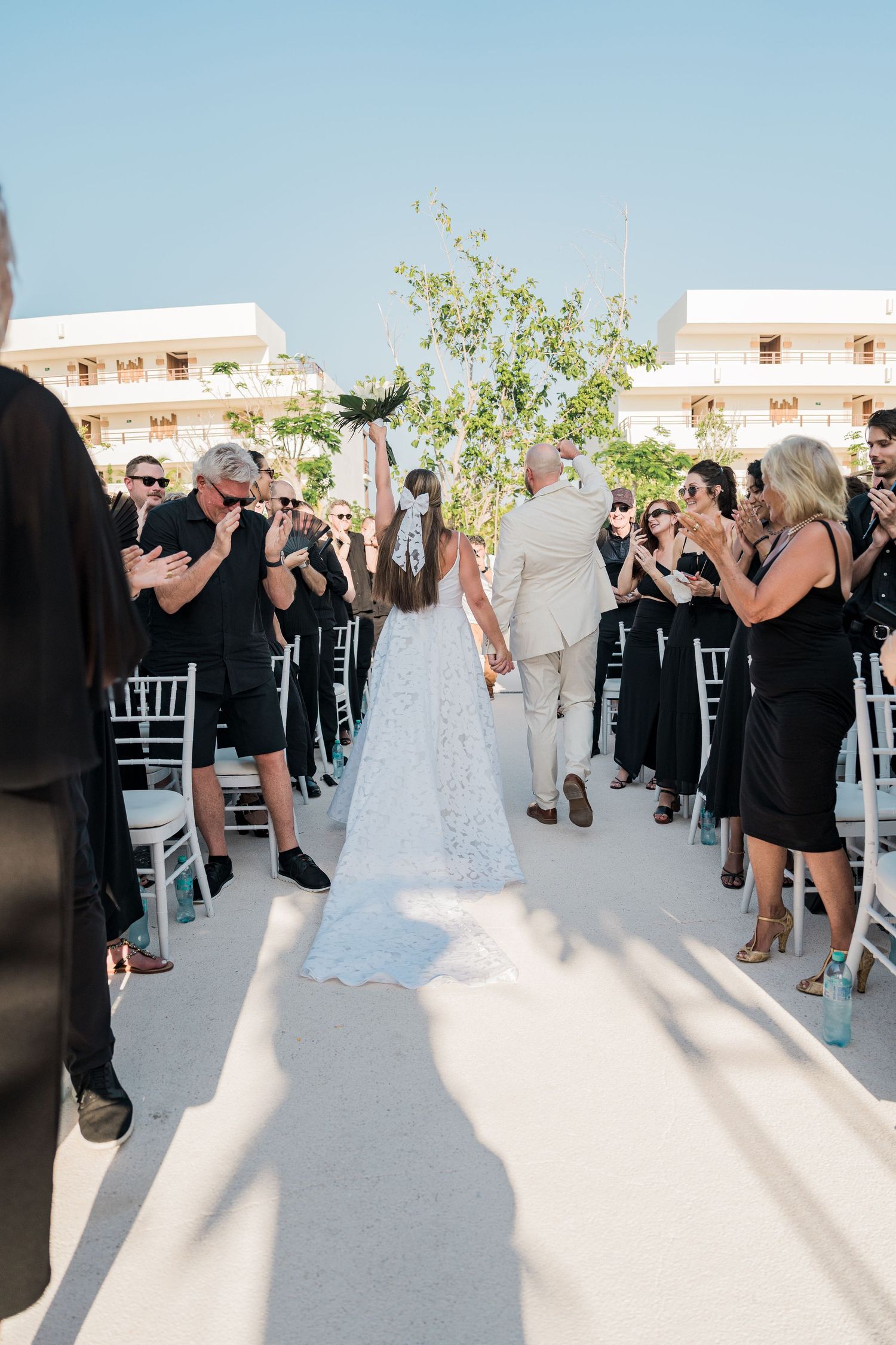 Bride and groom walk down the aisle at an outdoor wedding ceremony while guests in black attire applaud.