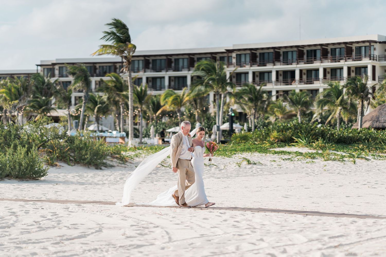 Couple shares romantic kiss on white sand beach with palm trees and luxury resort hotel backdrop on their wedding day.