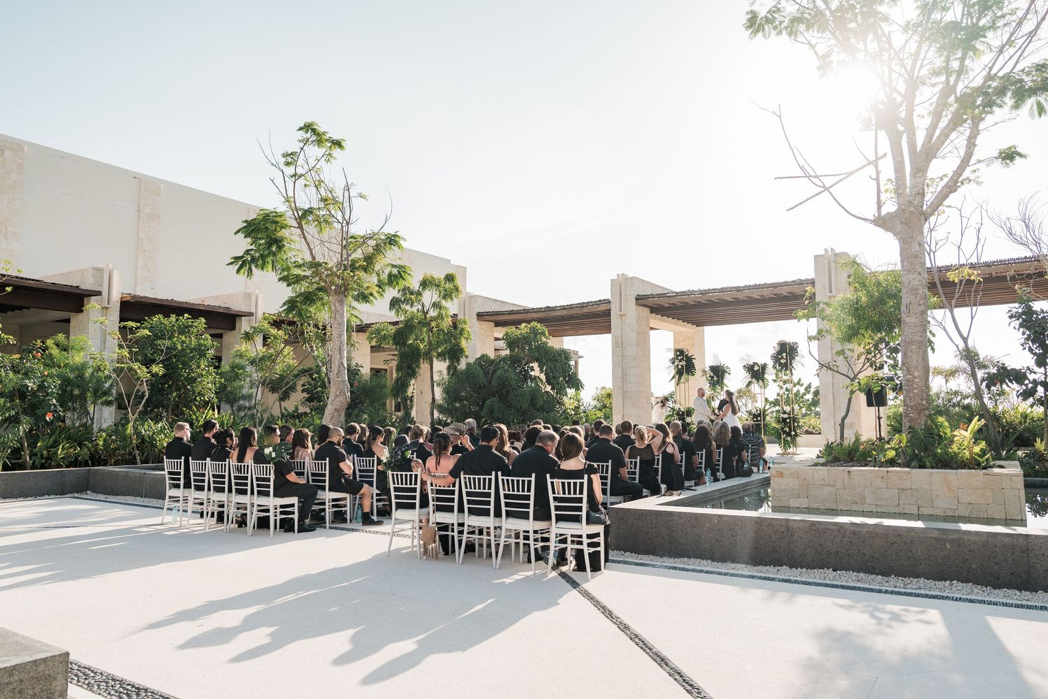 Outdoor wedding ceremony with guests seated in white chairs on a modern patio surrounded by tropical trees and concrete architecture.