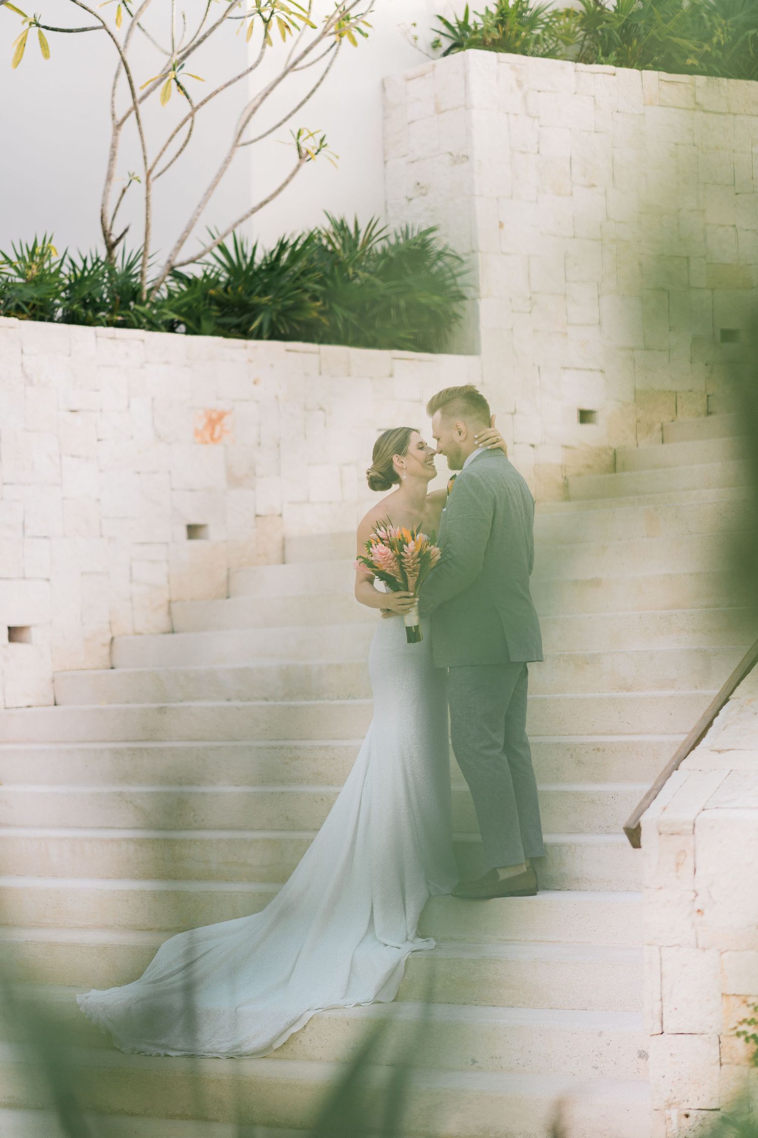Bride and groom share a romantic moment on elegant stone steps, bride in a flowing white gown holding a colorful bouquet.