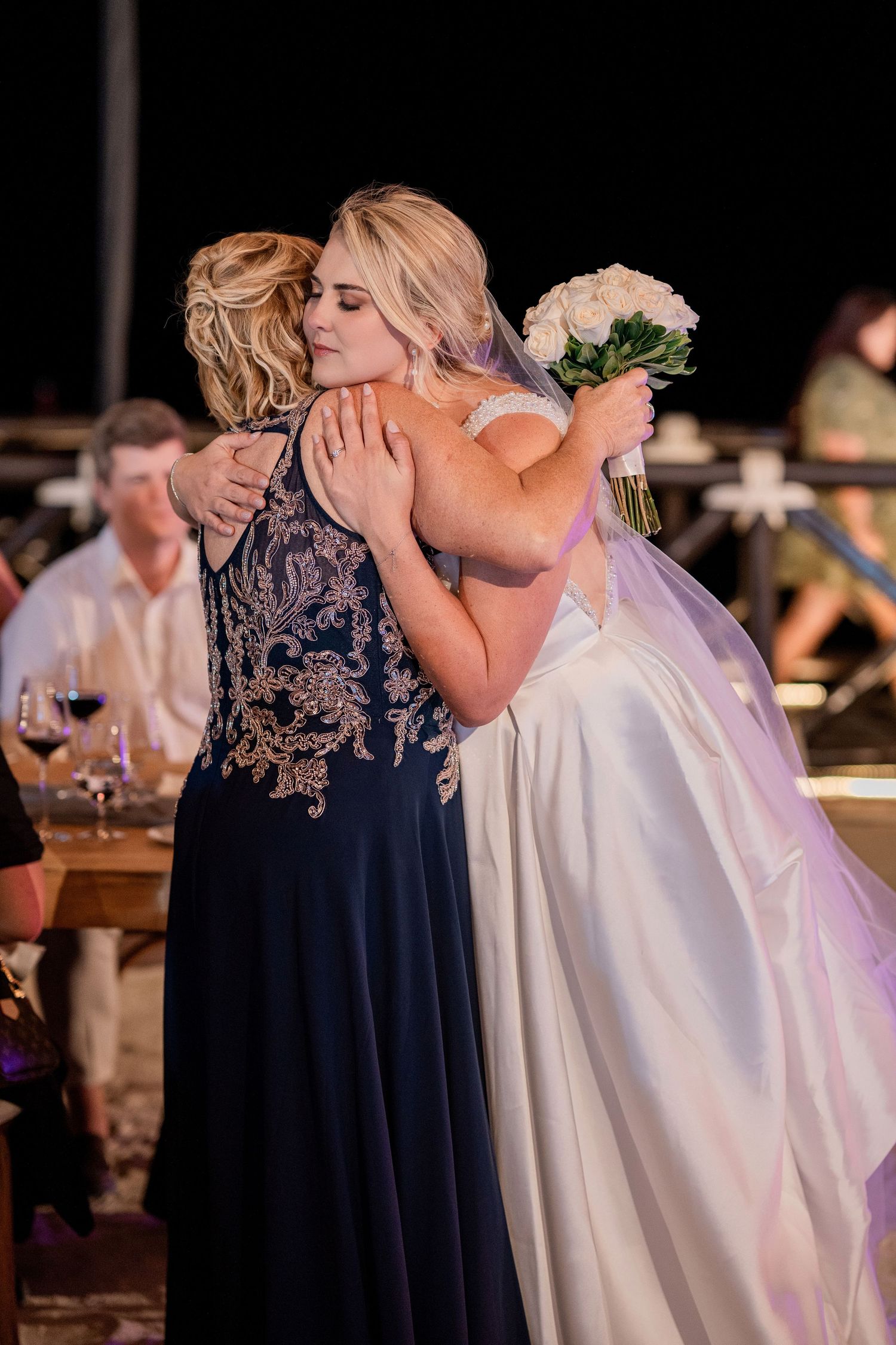 Bride in white gown hugging a woman in navy blue dress at an outdoor wedding reception, holding white bouquet.