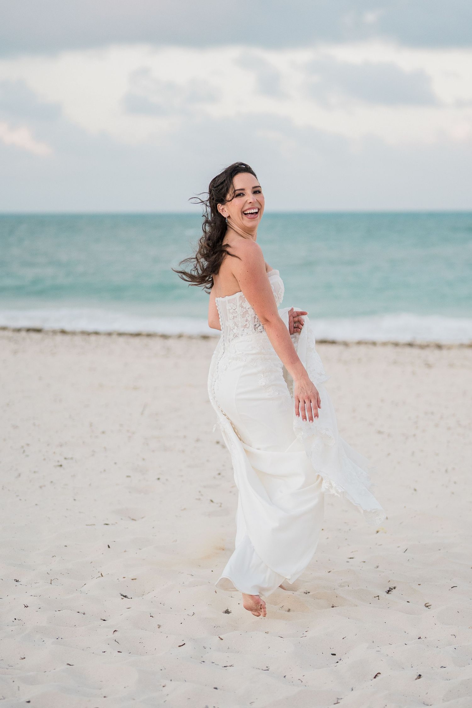 Laughing bride in white strapless gown walks barefoot on sandy beach with turquoise ocean waves in background.