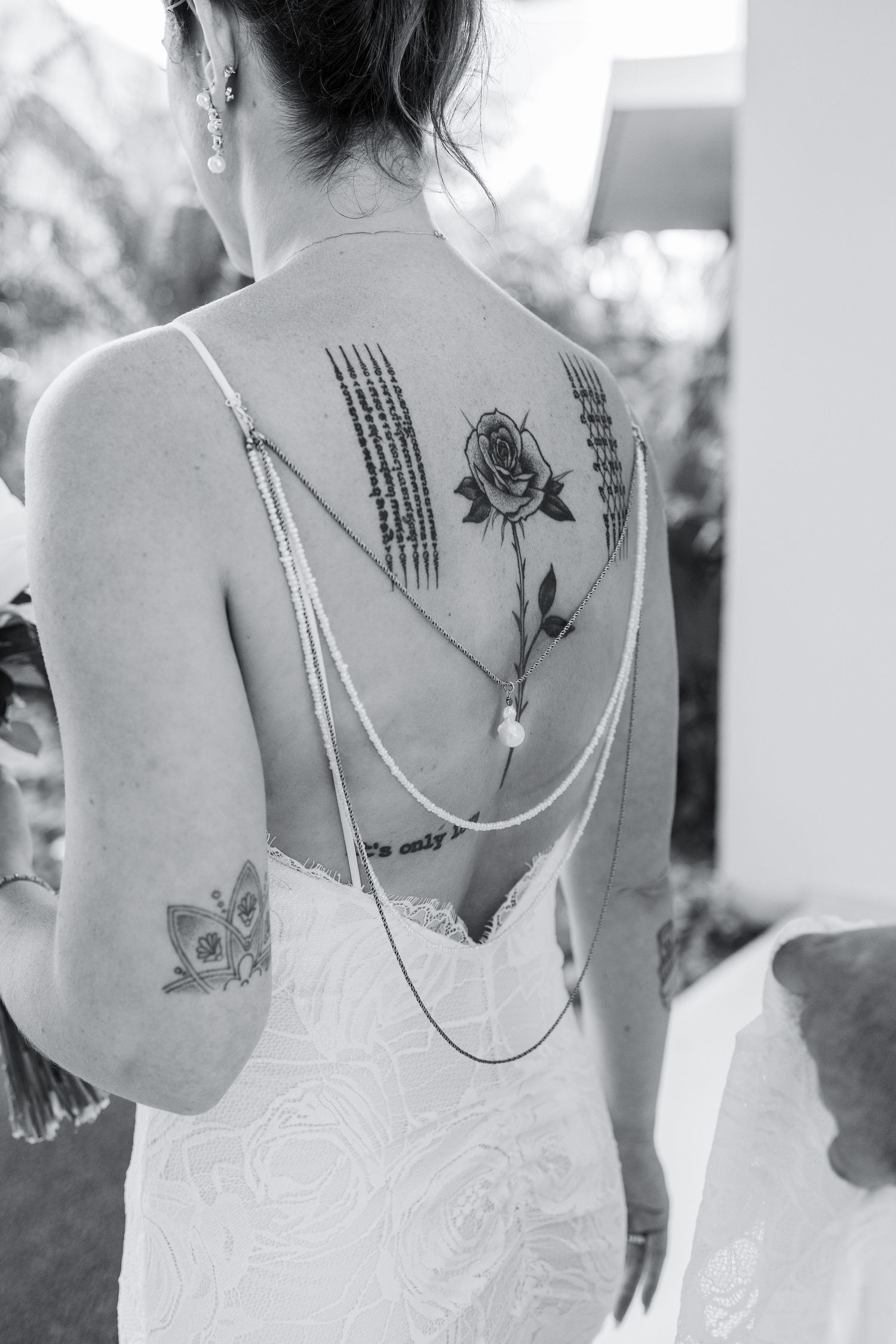 Black and white photo of bride in backless wedding dress with rose and script tattoos, wearing delicate chain back jewelry.