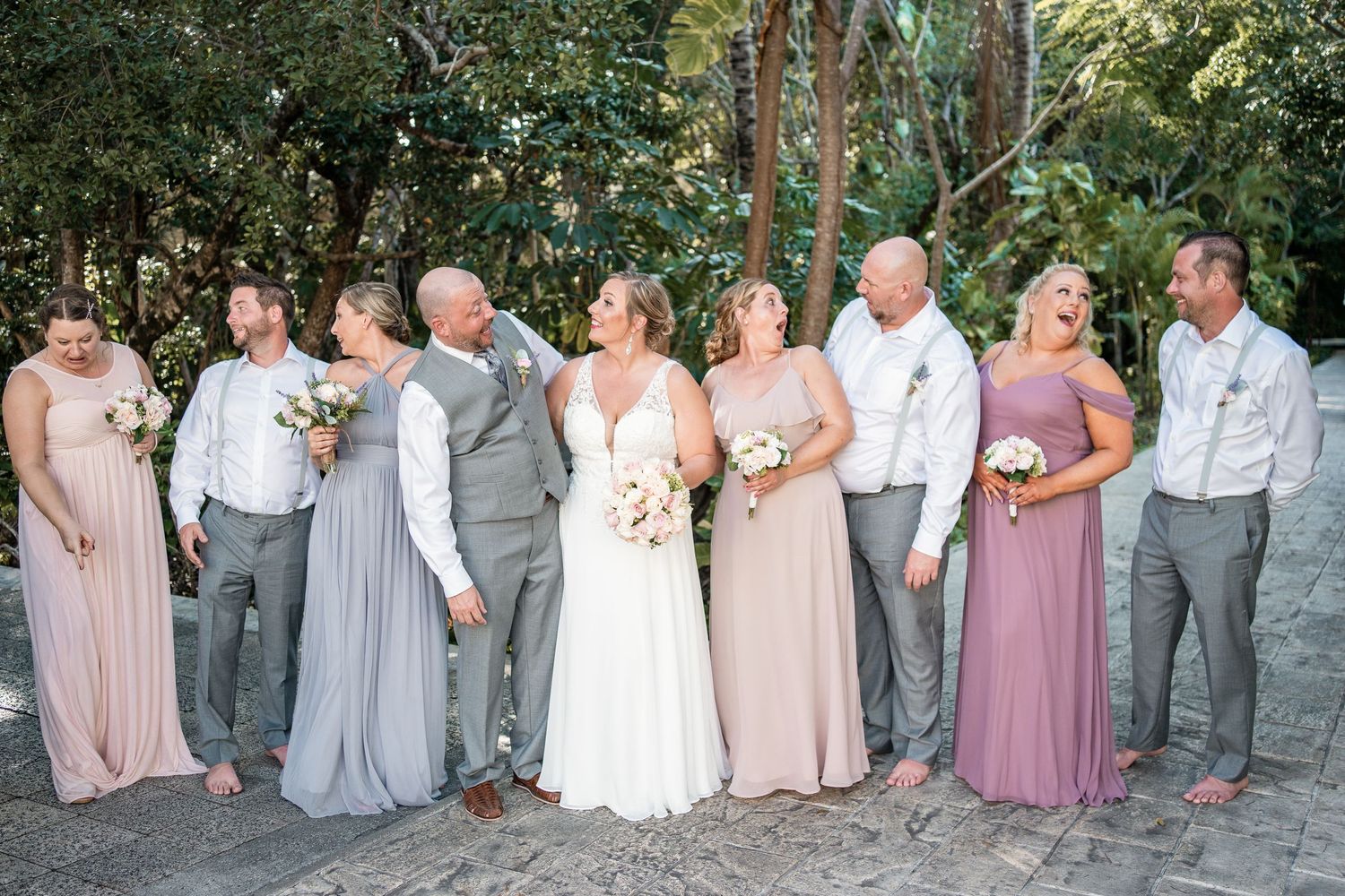 Full wedding party poses together on sandy beach path with tropical trees in background.