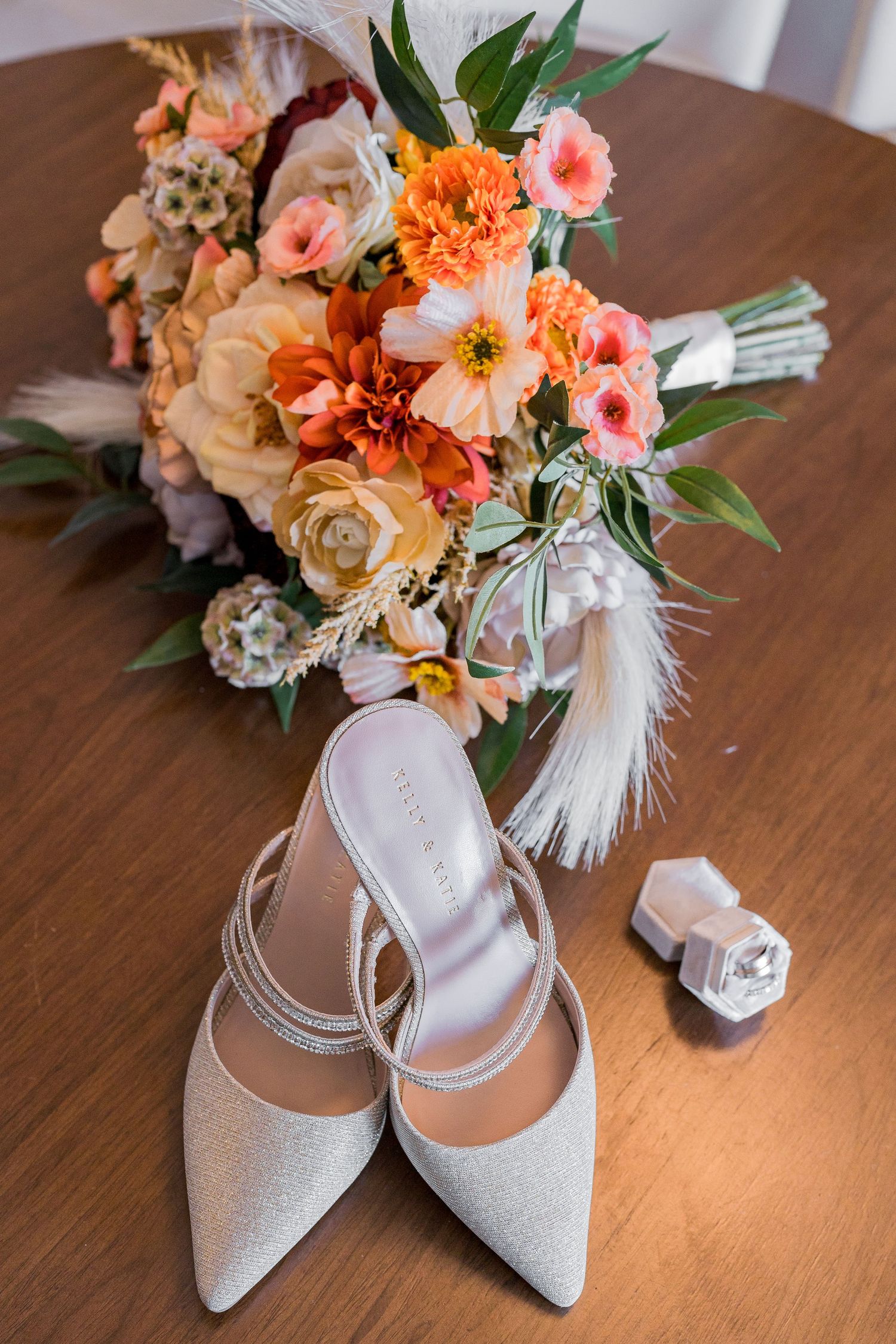 Elegant bridal flat shoes beside a vibrant orange and pink wedding bouquet with feathers and a ring box.