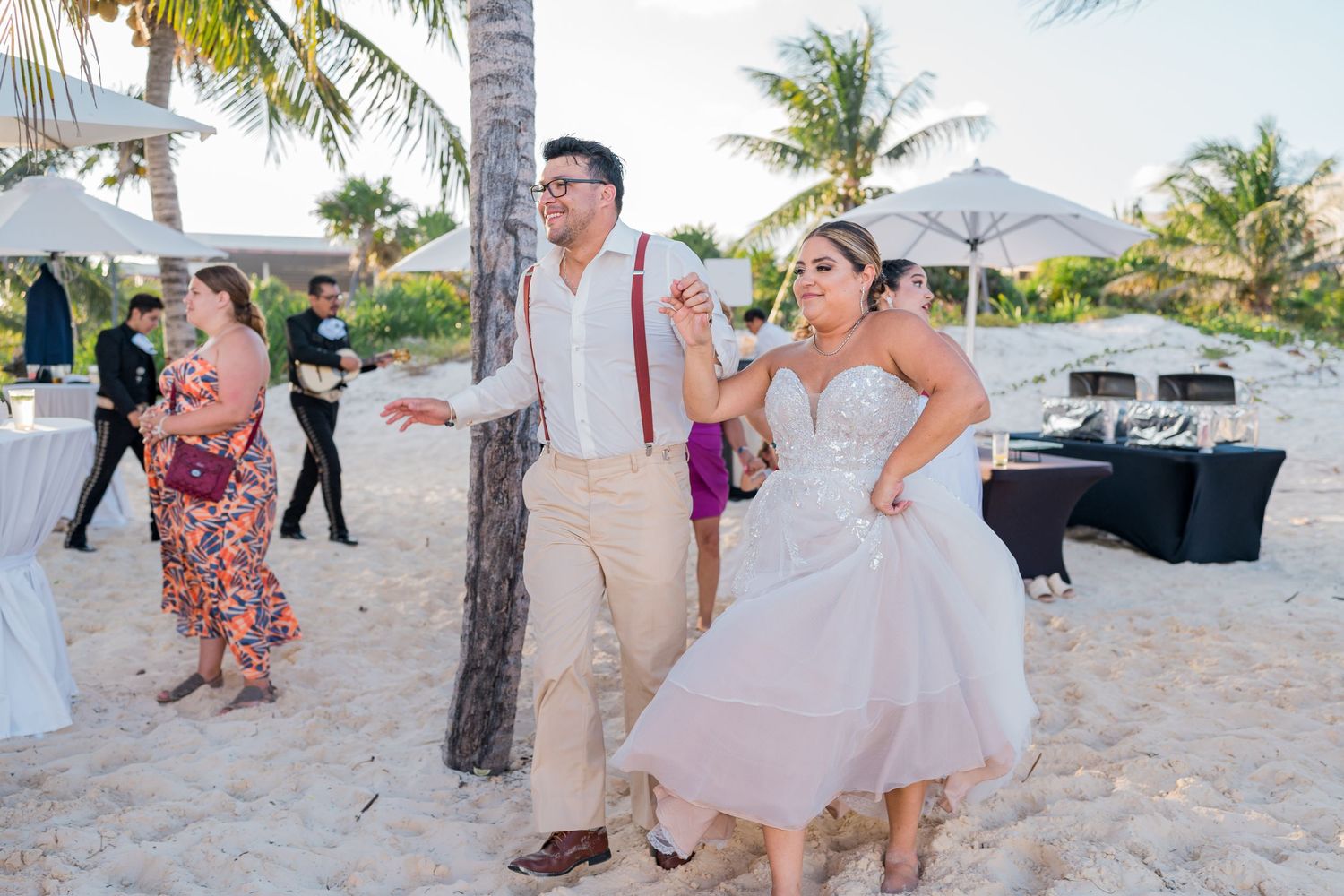Bride in short wedding dress dances with groom in suspenders on sandy beach at outdoor wedding reception.