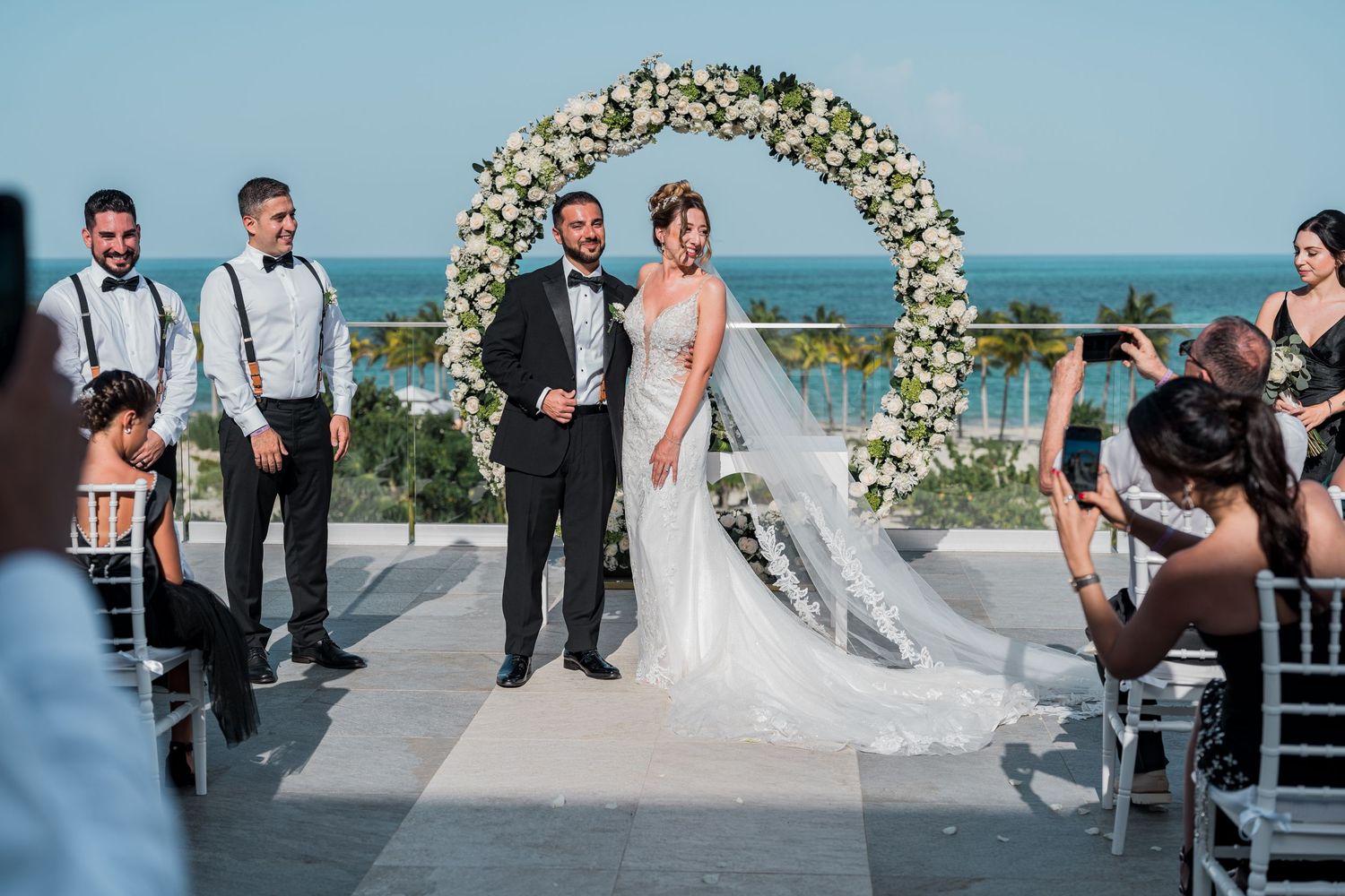 Bride and groom posing under a circular floral arch at an elegant oceanfront wedding ceremony.
